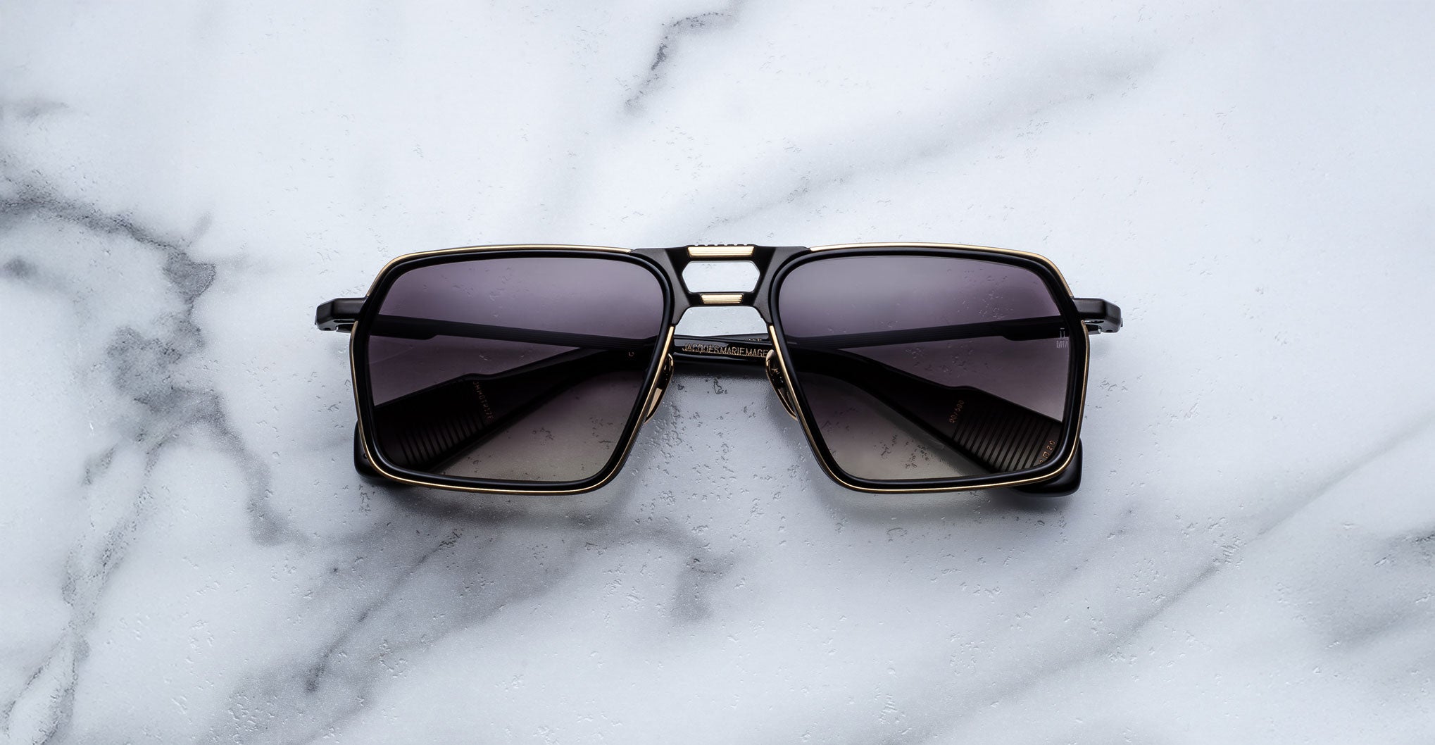 A pair of Jacques Marie Mage GT-R black square-framed sunglasses with dark tinted lenses rests on a white marble surface.