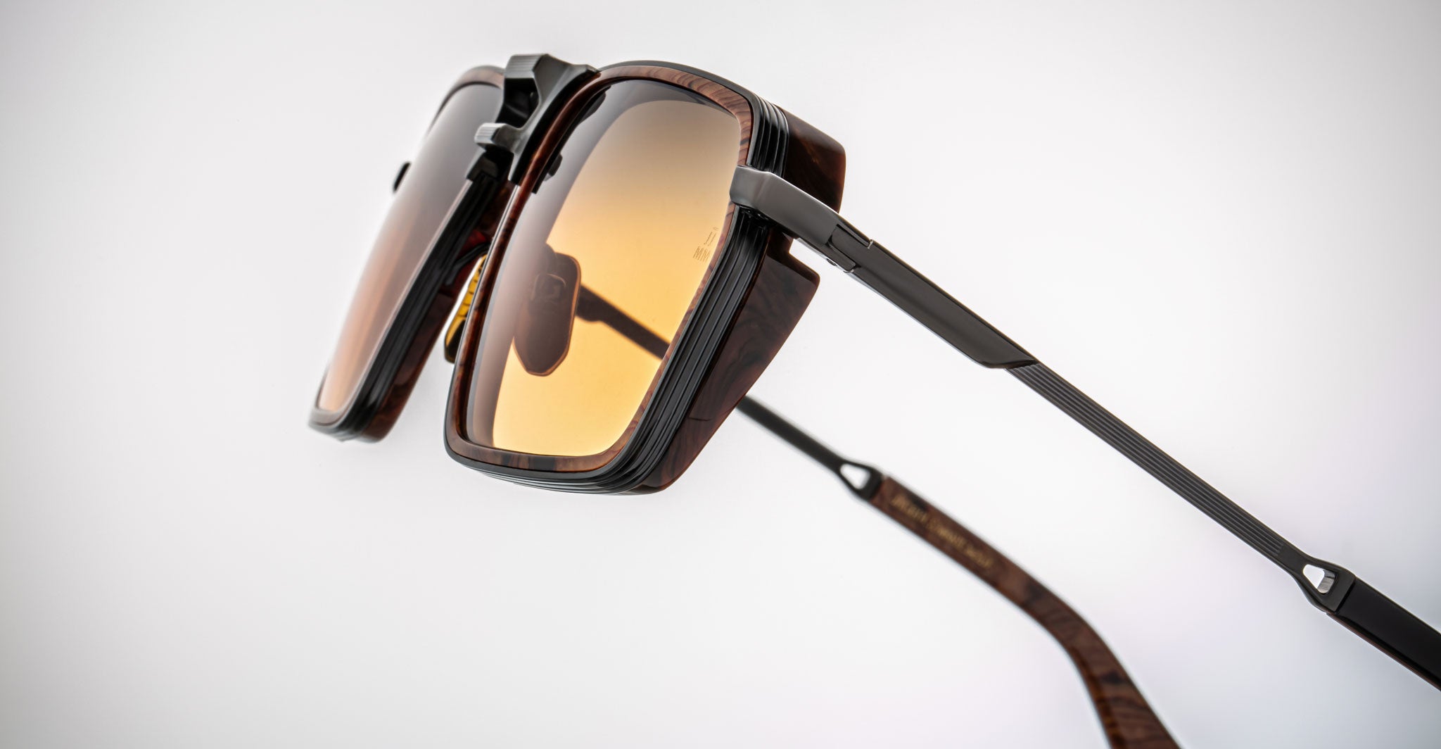 Close-up of Jacques Marie Mage GT-R sunglasses with dark brown frames, amber-tinted lenses, thin black metal arms, and a modern geometric design, set against a plain white background.
