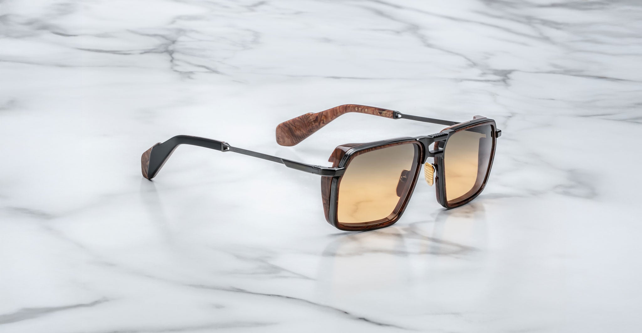 A pair of Jacques Marie Mage GT-R sunglasses featuring brown square frames, gradient amber lenses, and wood-textured arms, displayed on a white marble surface.