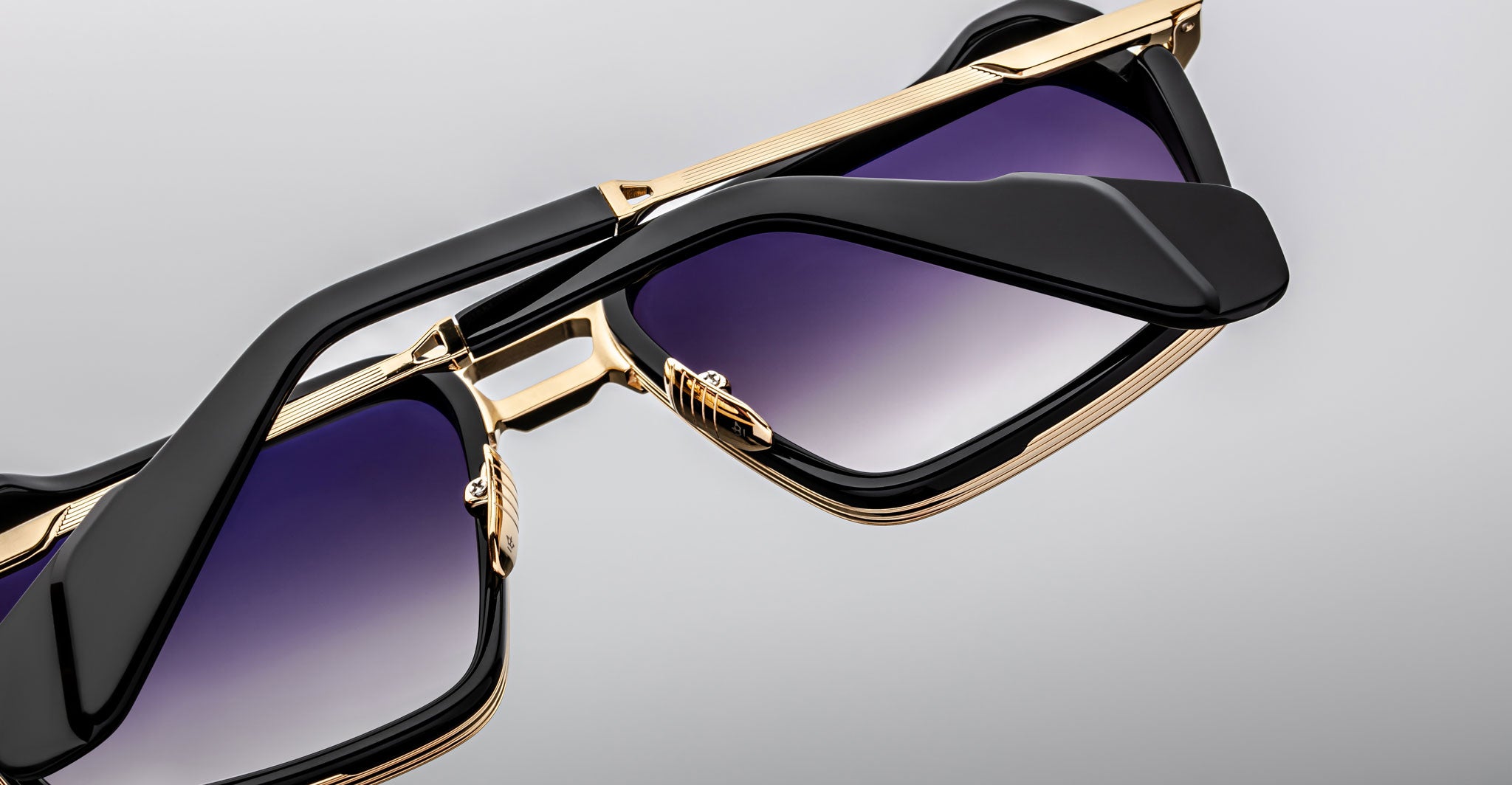 Close-up of two Jacques Marie Mage GT-R sunglasses with gold frames, black arms, and gradient purple lenses, one stacked atop the other against a plain grey background.