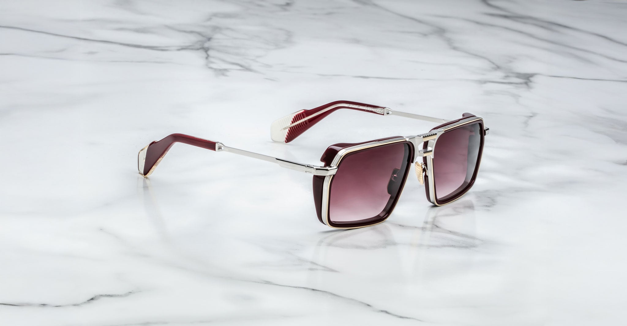 Jacques Marie Mage GT-R sunglasses with square frames, dark gradient lenses, maroon arms, and metallic accents are displayed on a white marble surface.
