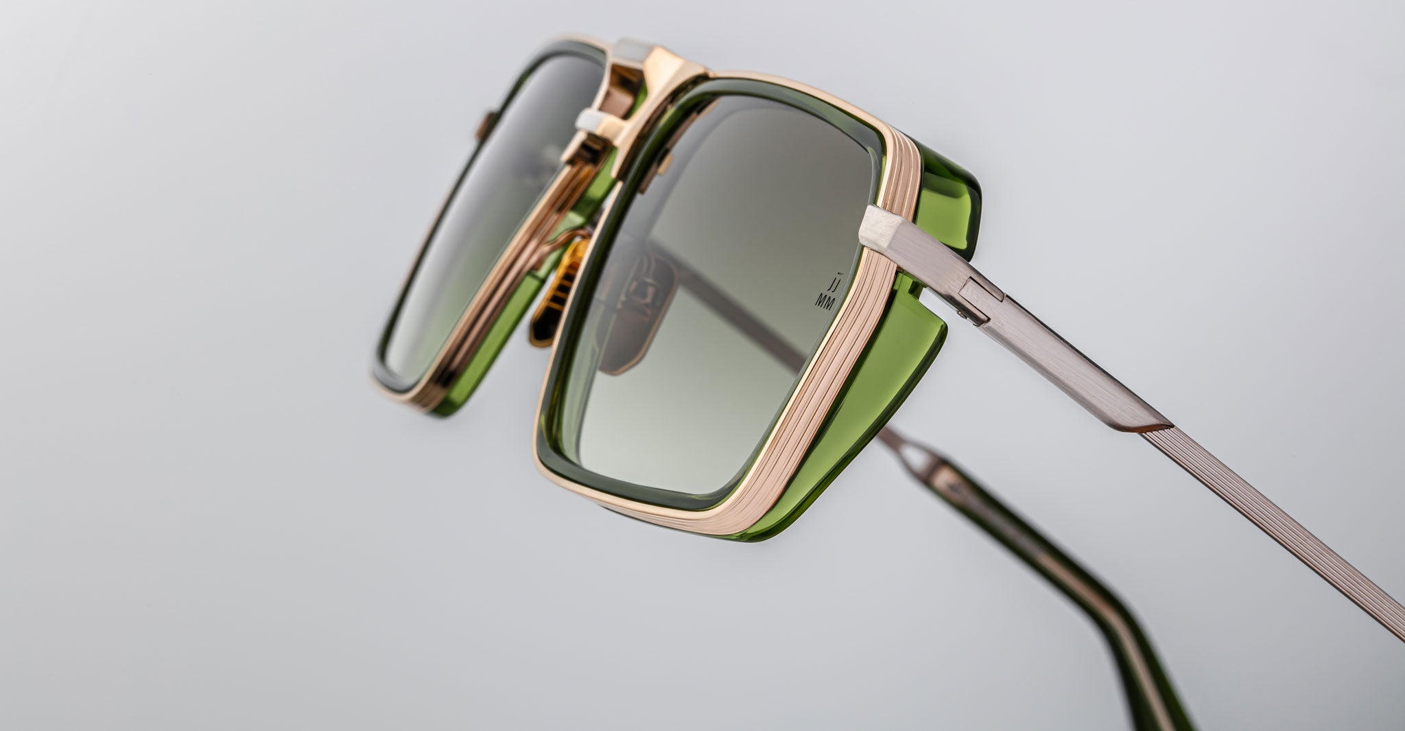 Close-up of Jacques Marie Mage GT-R sunglasses featuring square frames, green-tinted lenses, gold and green accents, and metal arms, set against a plain light gray background.