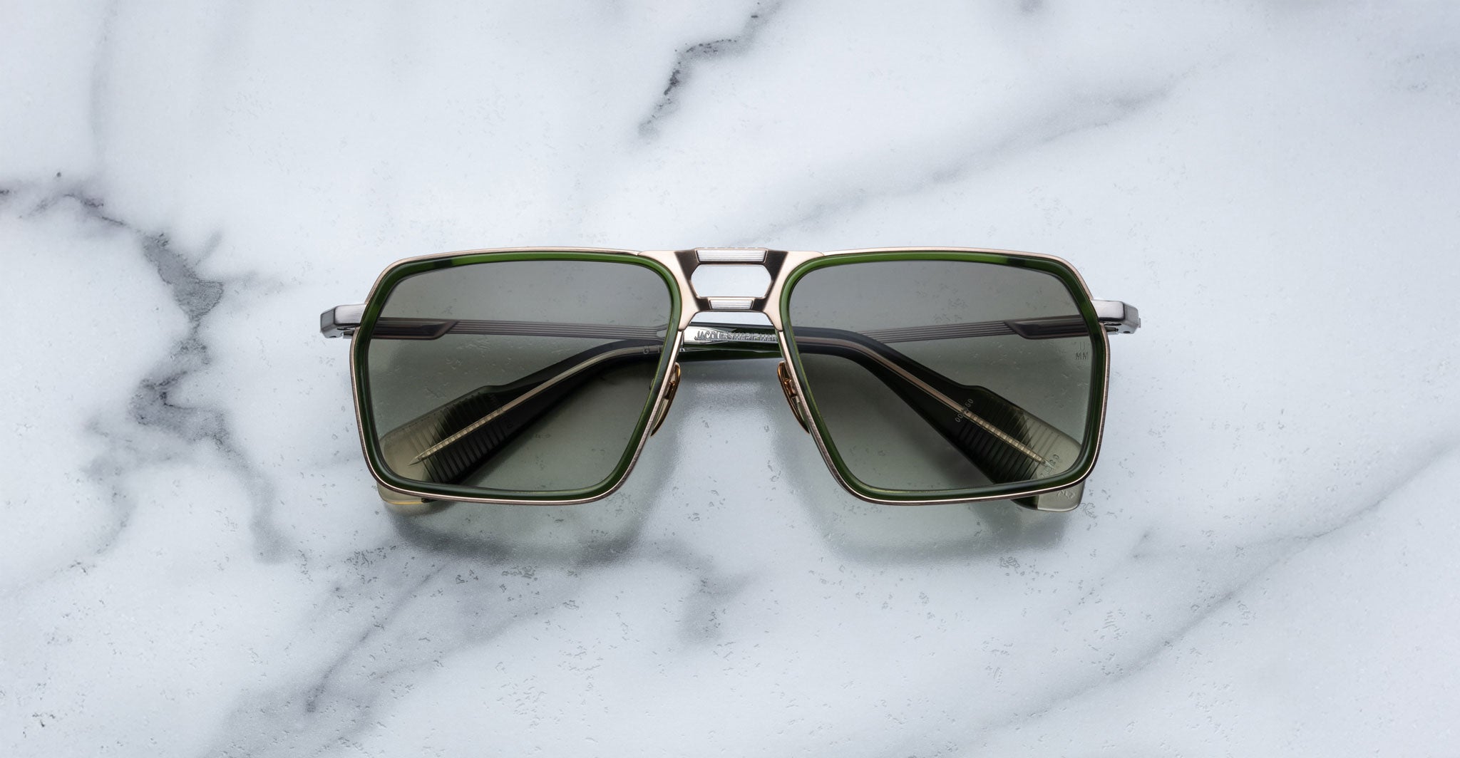 The Jacques Marie Mage GT-R, featuring square-shaped sunglasses with green-tinted lenses and metallic frames, is displayed on a white marble surface.