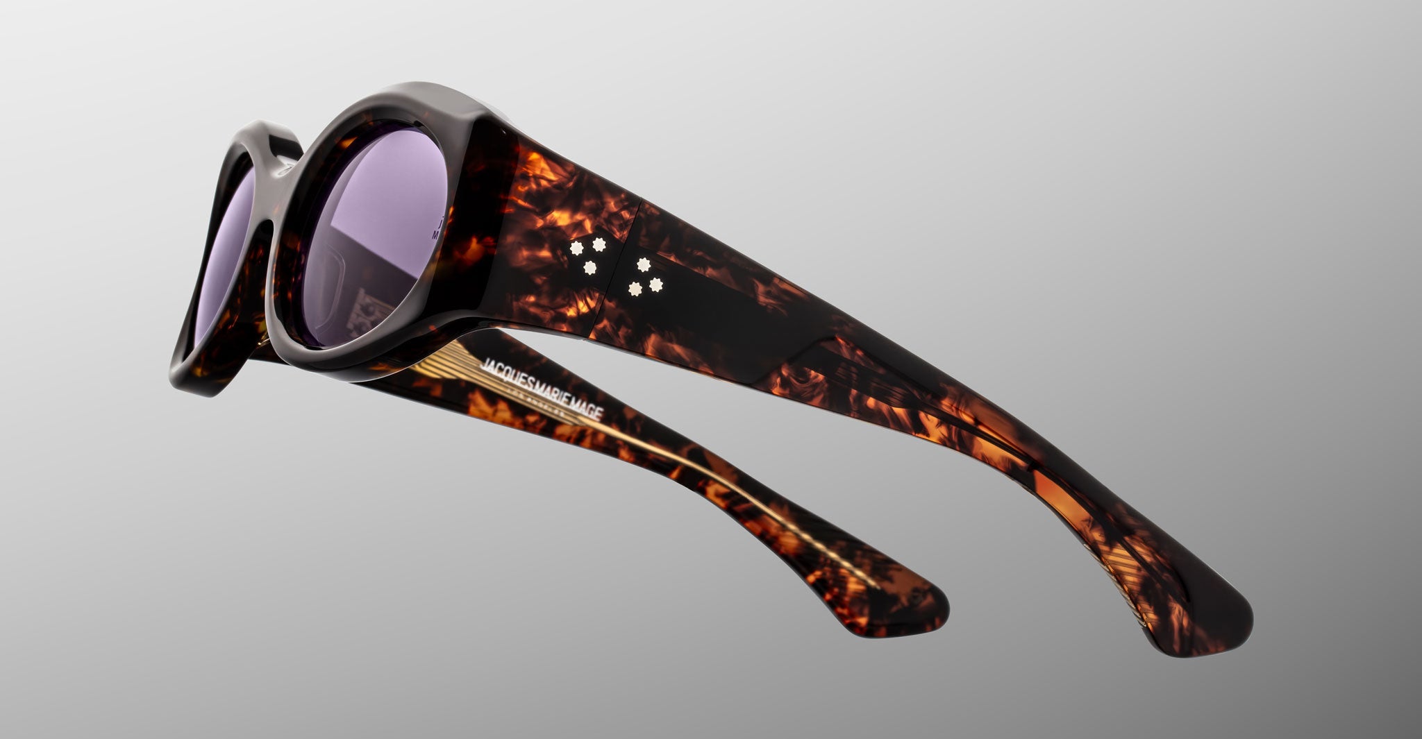 The Jacques Marie Mage Gia sunglasses, handcrafted in Japan, showcase thick tortoiseshell frames and dark tinted lenses by Jacques Marie Mage, set against a plain grey background.