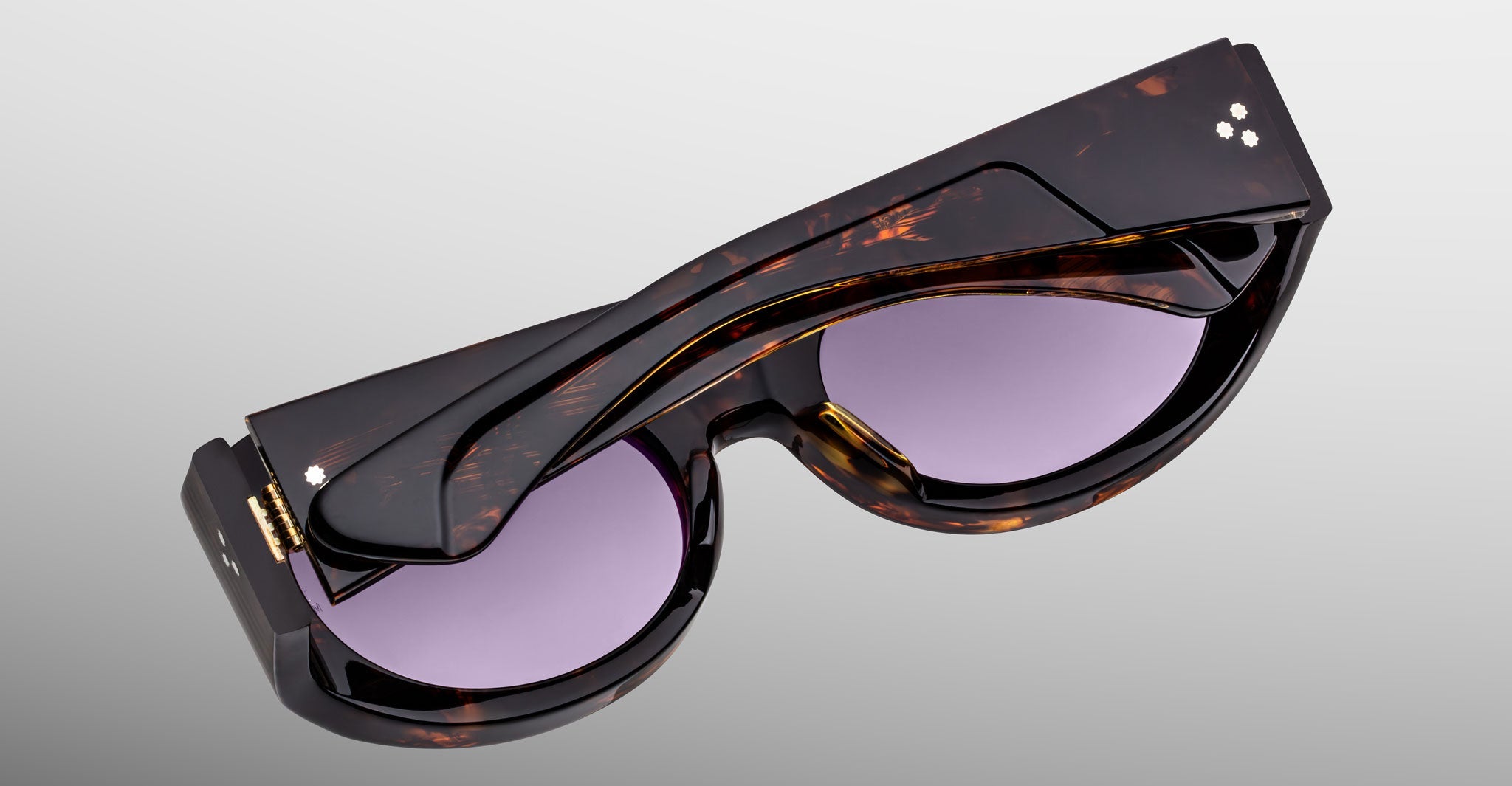 The Jacques Marie Mage Gia sunglasses, handcrafted in Japan with an oval acetate frame and purple lenses, are neatly folded and displayed against a plain grey background.
