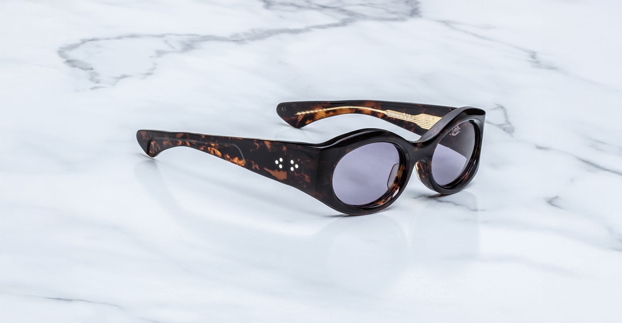 A pair of Jacques Marie Mage Gia oval acetate sunglasses by Jacques Marie Mage, handcrafted in Japan with tortoiseshell frames and round dark lenses, displayed on a white marble surface.
