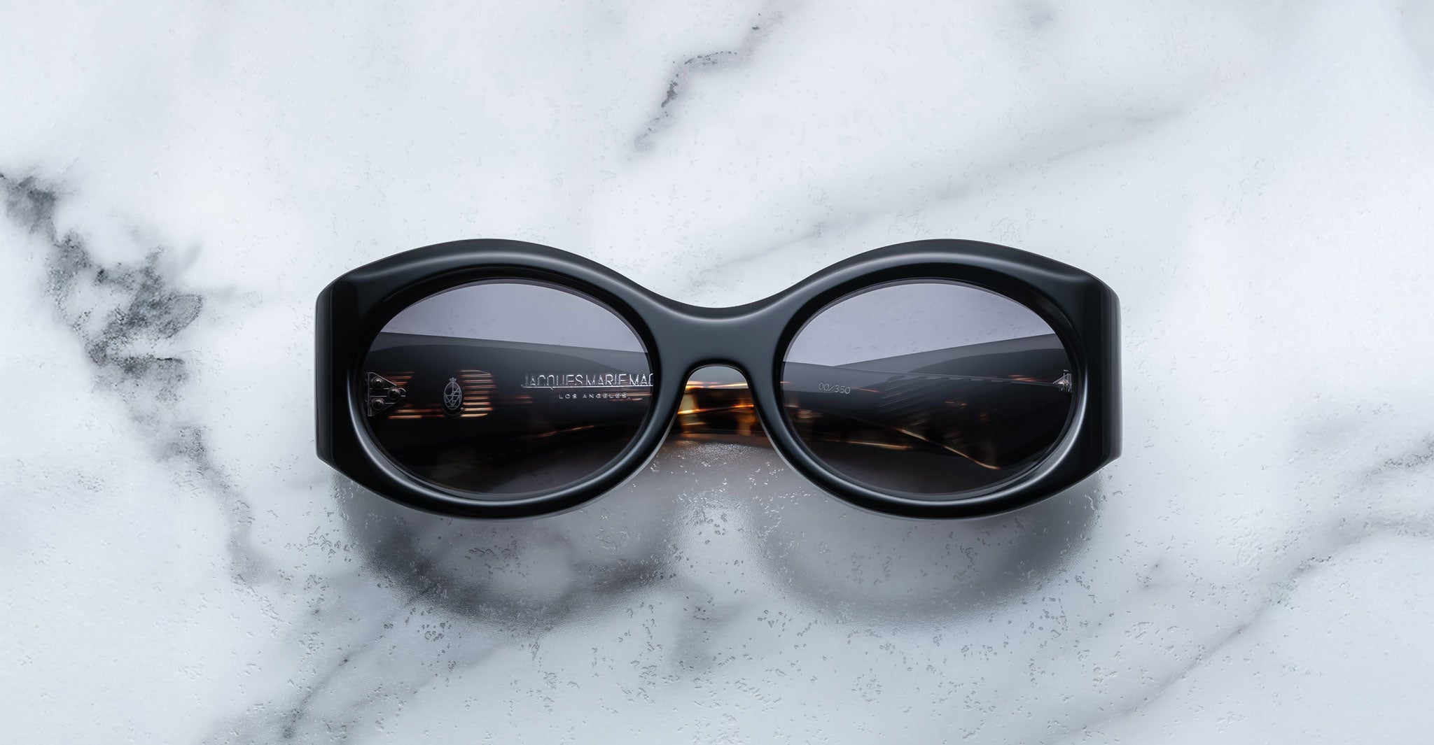 A pair of Jacques Marie Mage Gia black, thick-framed sunglasses with grey lenses rests on a white marble surface with grey veins, with faint brand details visible on the inside of the left lens.