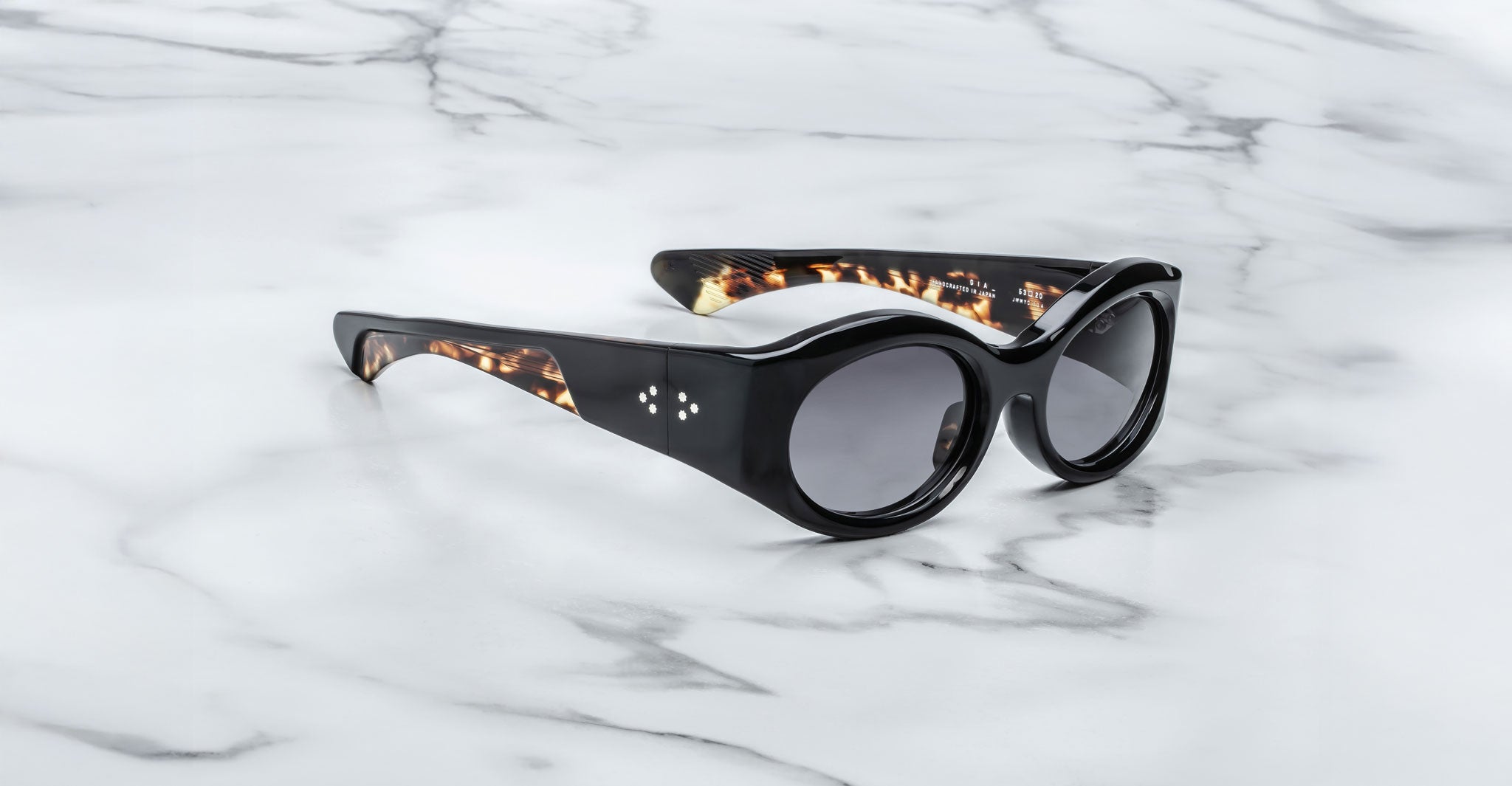 A pair of Jacques Marie Mage Gia black color, thick-framed sunglasses with grey lenses sits on a white marble surface with grey veins, with faint brand details visible on the inside of the left lens.
