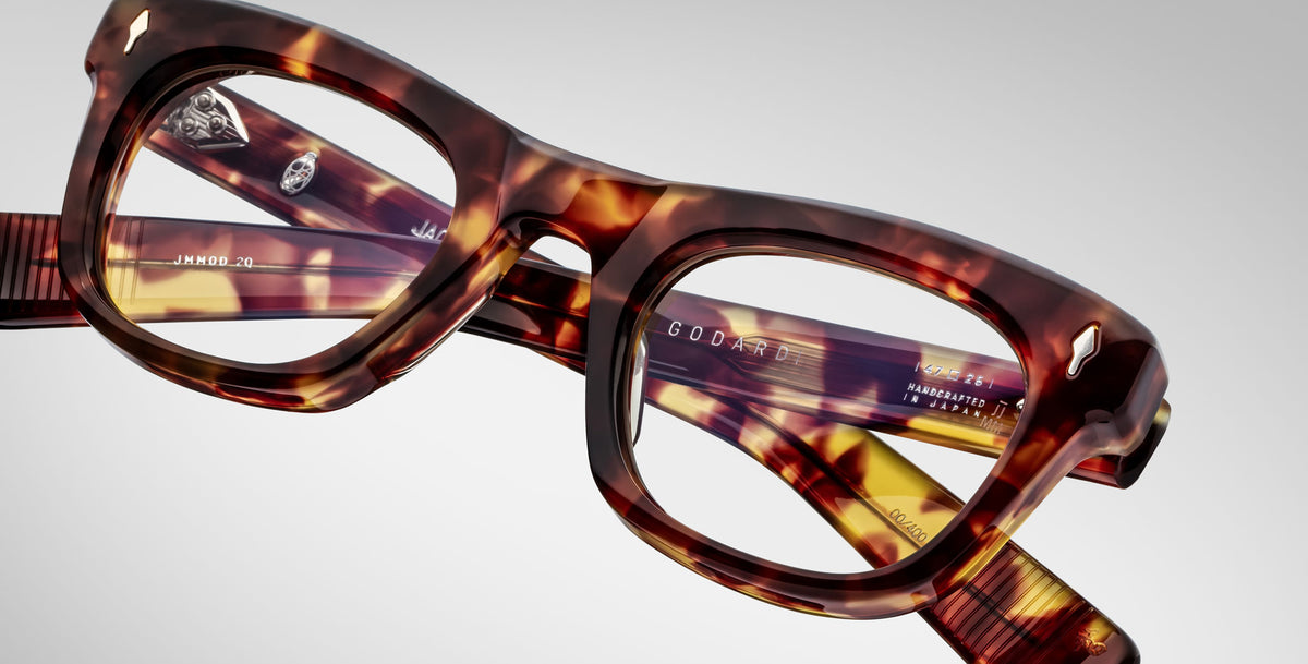 Close-up of Jacques Marie Mage Godard tortoiseshell eyeglasses with thick rectangular frames, resting on their side. Product details are visible on the inner arms against a light gray background.