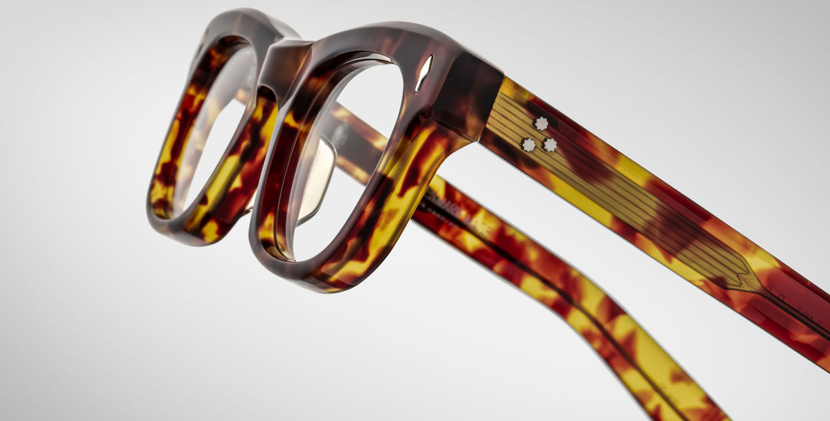 Close-up of Jacques Marie Mage Godard eyeglasses featuring thick, brown and yellow tortoiseshell-patterned plastic frames, shown at an angle highlighting the front and side arms against a plain light background.