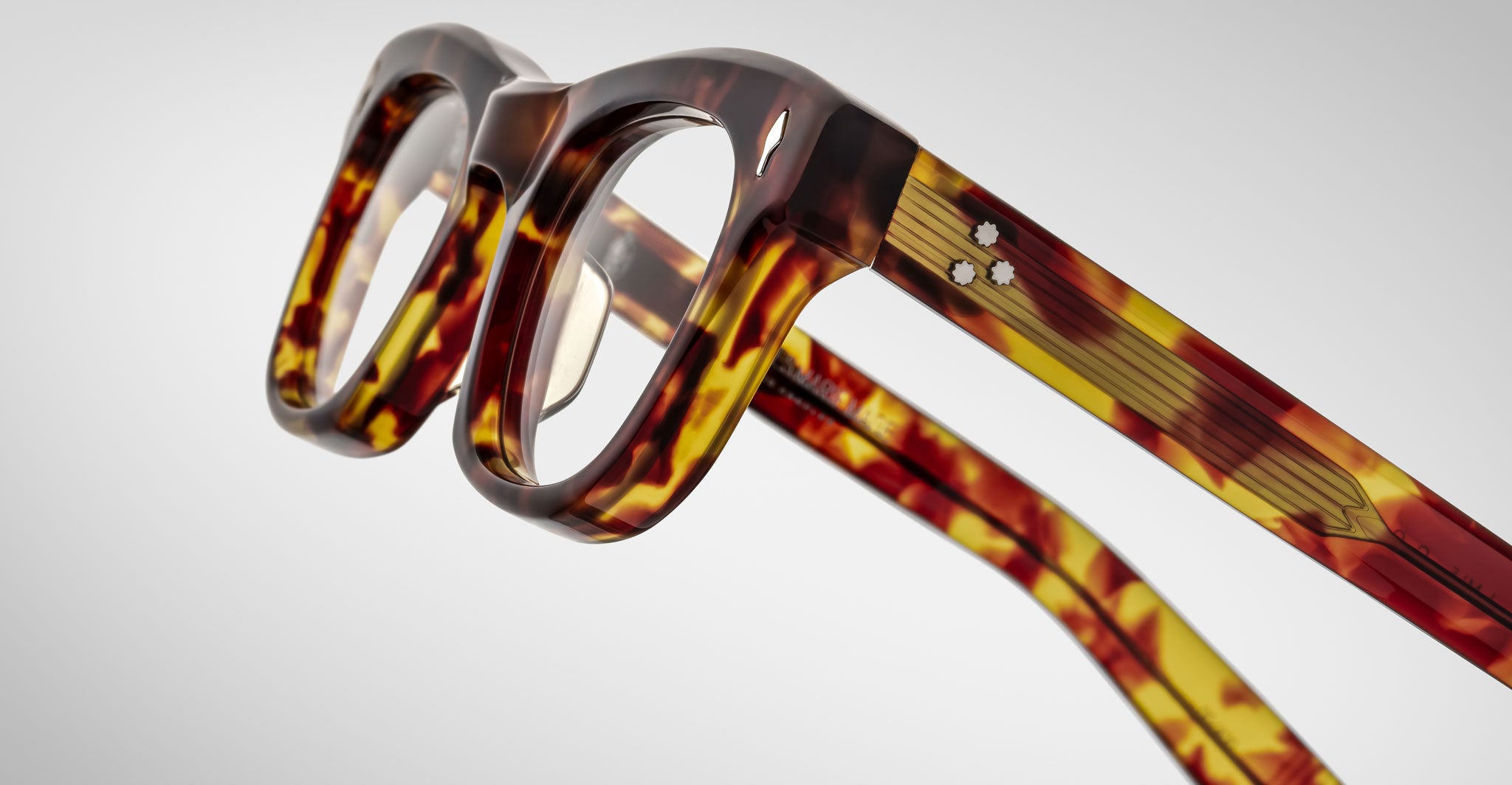 Close-up of Jacques Marie Mage Godard eyeglasses featuring thick, brown and yellow tortoiseshell-patterned plastic frames, shown at an angle highlighting the front and side arms against a plain light background.