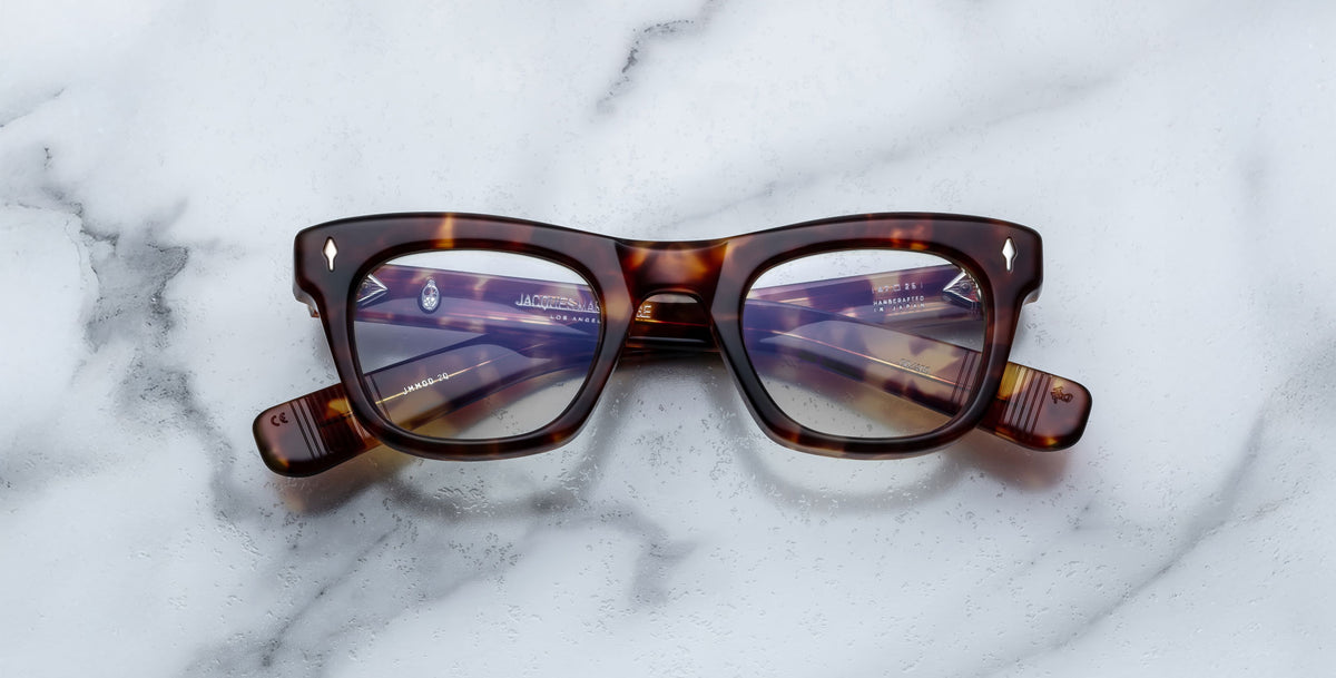 A pair of Jacques Marie Mage Godard tortoiseshell rectangular eyeglasses with clear lenses rests on a white marble surface. The arms are folded, showing text on the inside of the left arm.
