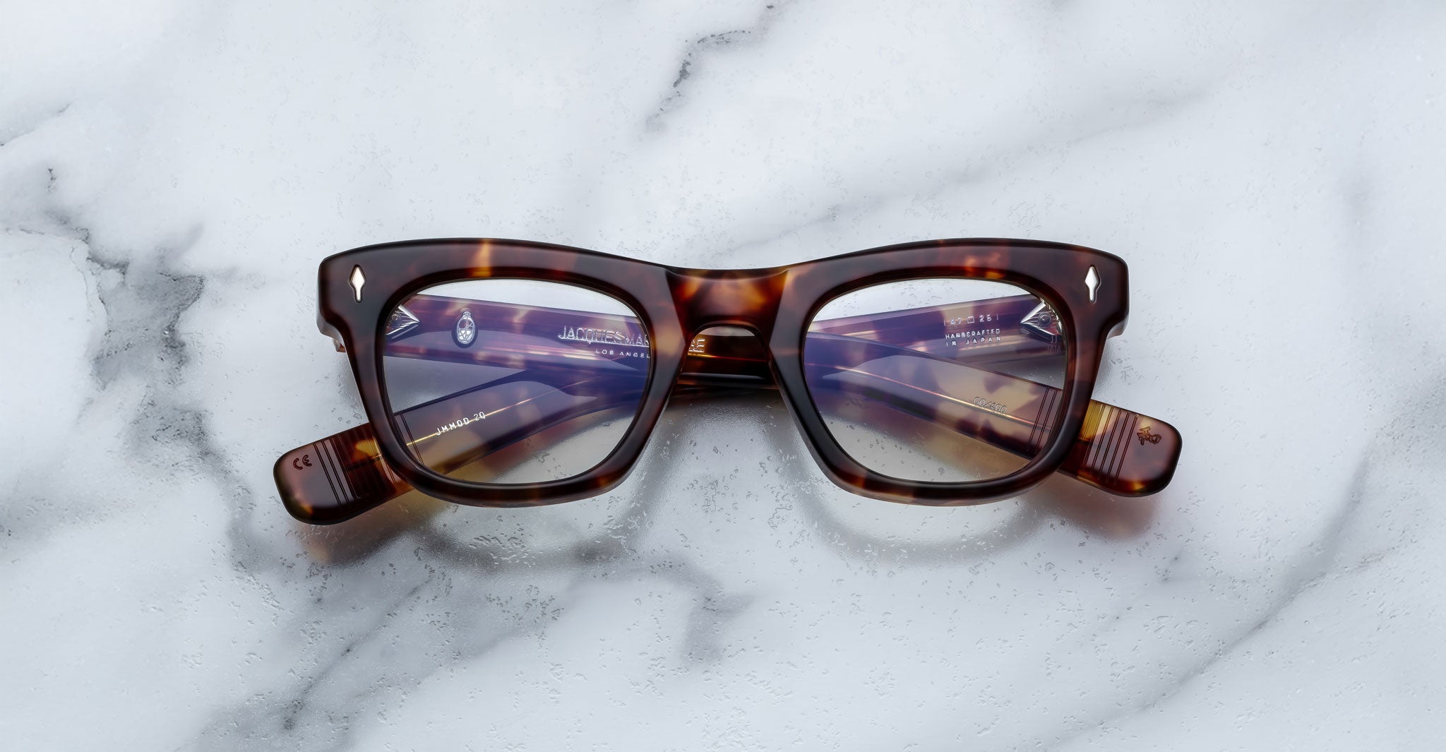 A pair of Jacques Marie Mage Godard tortoiseshell rectangular eyeglasses with clear lenses rests on a white marble surface. The arms are folded, showing text on the inside of the left arm.