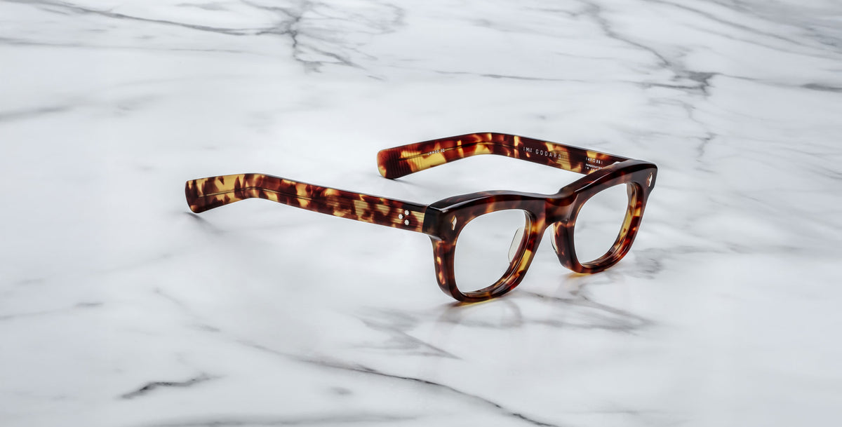 A pair of Jacques Marie Mage Godard thick-rimmed, tortoiseshell eyeglasses rests on a white marble surface with gray veining.