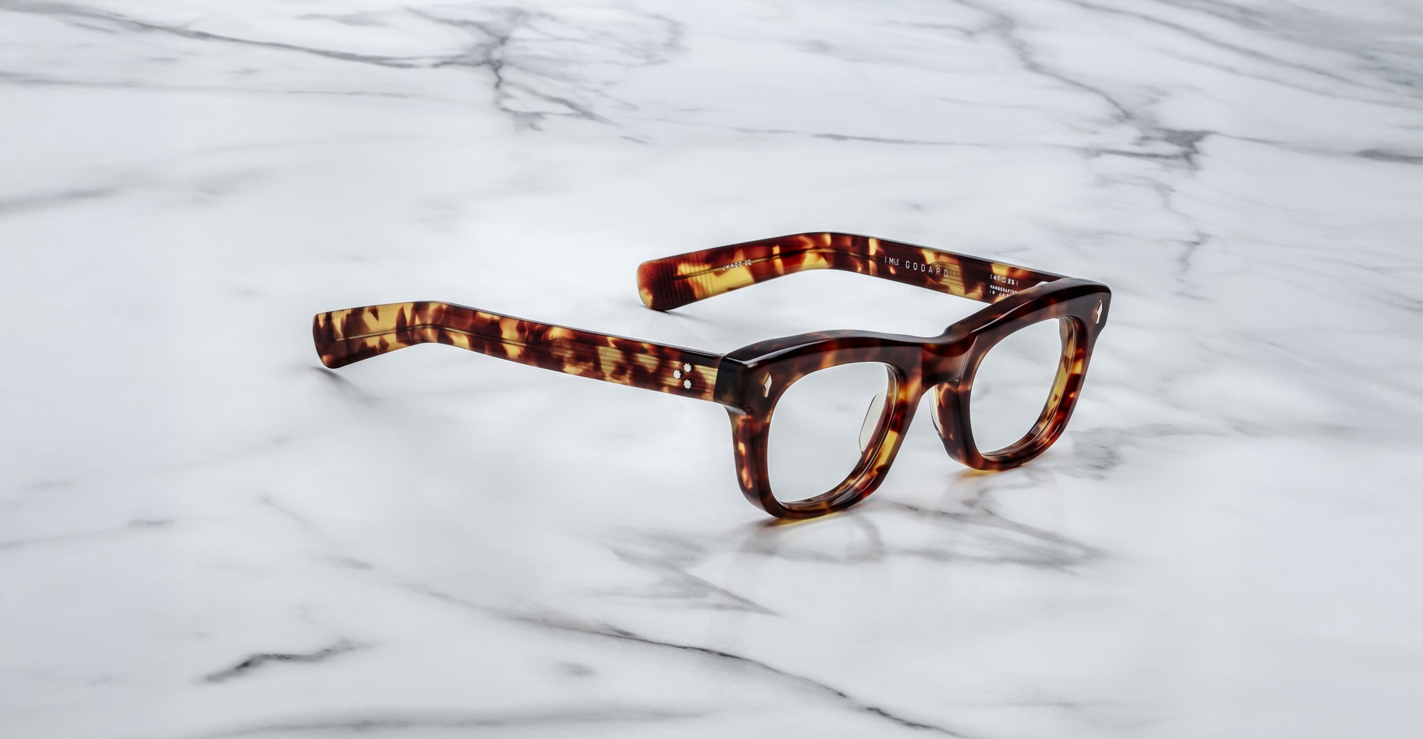 A pair of Jacques Marie Mage Godard thick-rimmed, tortoiseshell eyeglasses rests on a white marble surface with gray veining.