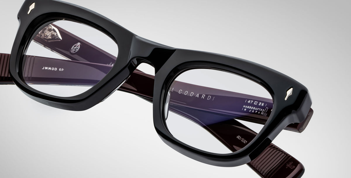 Close-up view of Jacques Marie Mage Godard black thick-rimmed eyeglasses with clear lenses. The inside arms show brand markings and model details. Set against a plain, light-colored background.