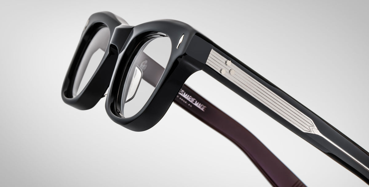A close-up of Jacques Marie Mage Godard thick-rimmed black eyeglasses with silver detailing along the temples, resting on a neutral gray background. White text is inscribed inside the arm.