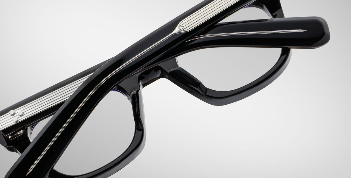 Close-up of Jacques Marie Mage Godard black eyeglasses with silver-detailed temples, folded against a plain light gray background.