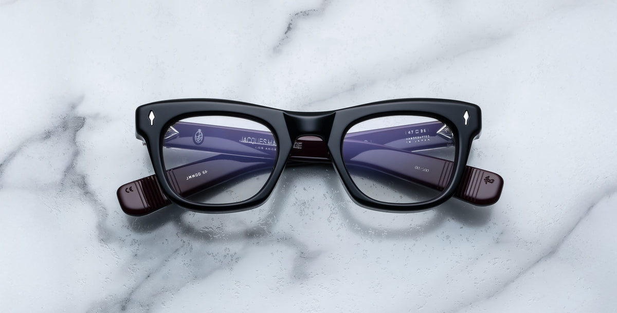 The Jacques Marie Mage Godard glasses feature a black rectangular frame with purple-tinted lenses and silver accents on the upper corners, shown on a white marble surface.