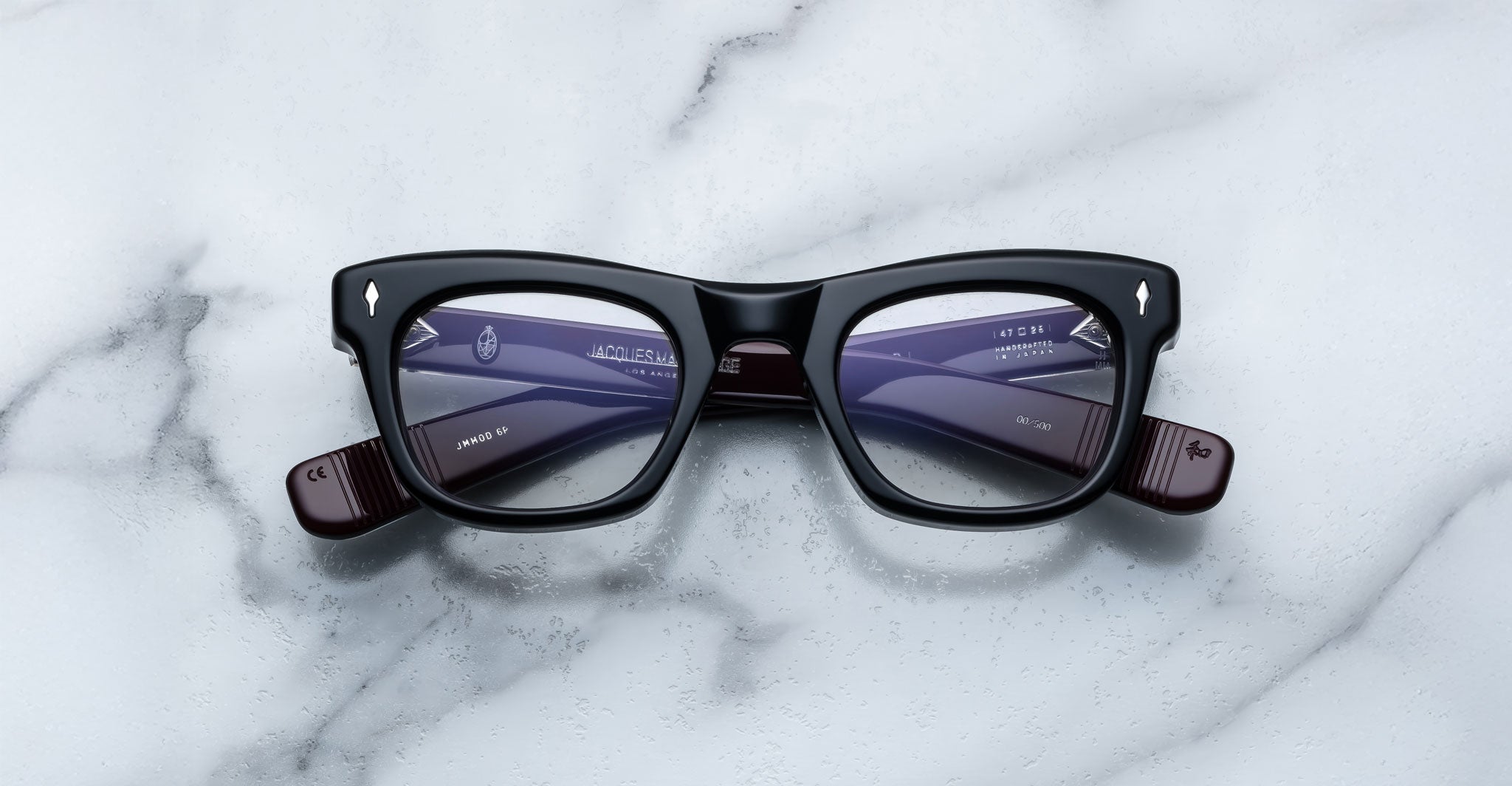 The Jacques Marie Mage Godard glasses feature a black rectangular frame with purple-tinted lenses and silver accents on the upper corners, shown on a white marble surface.