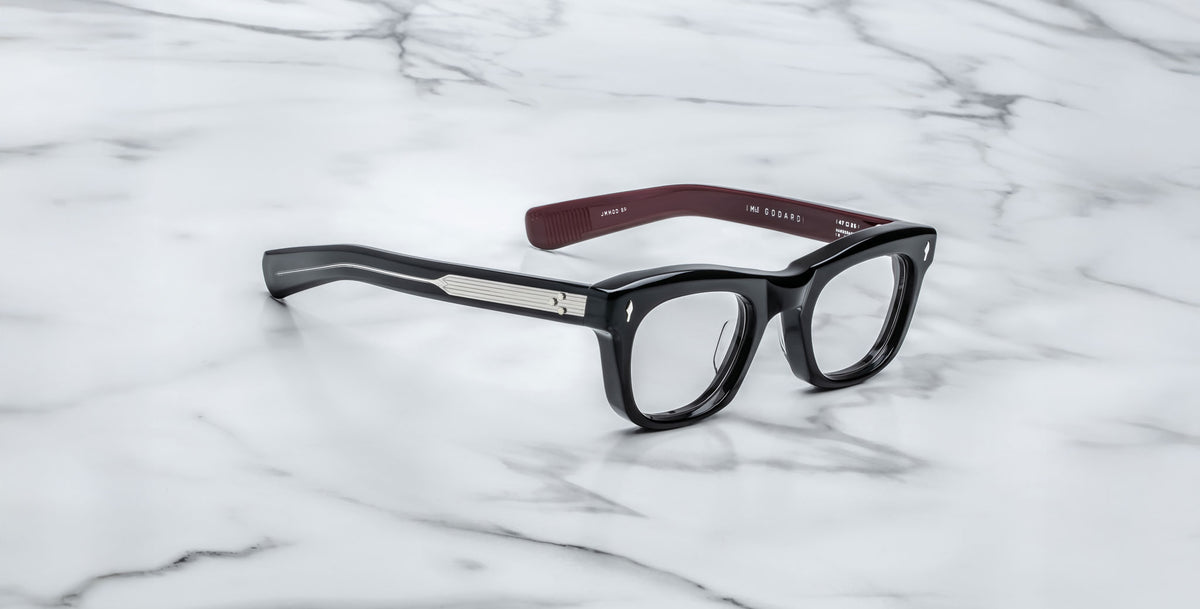 A pair of Jacques Marie Mage Godard black rectangular eyeglasses with thick frames, silver side accents, and brown temple interiors rests on a white marble surface.