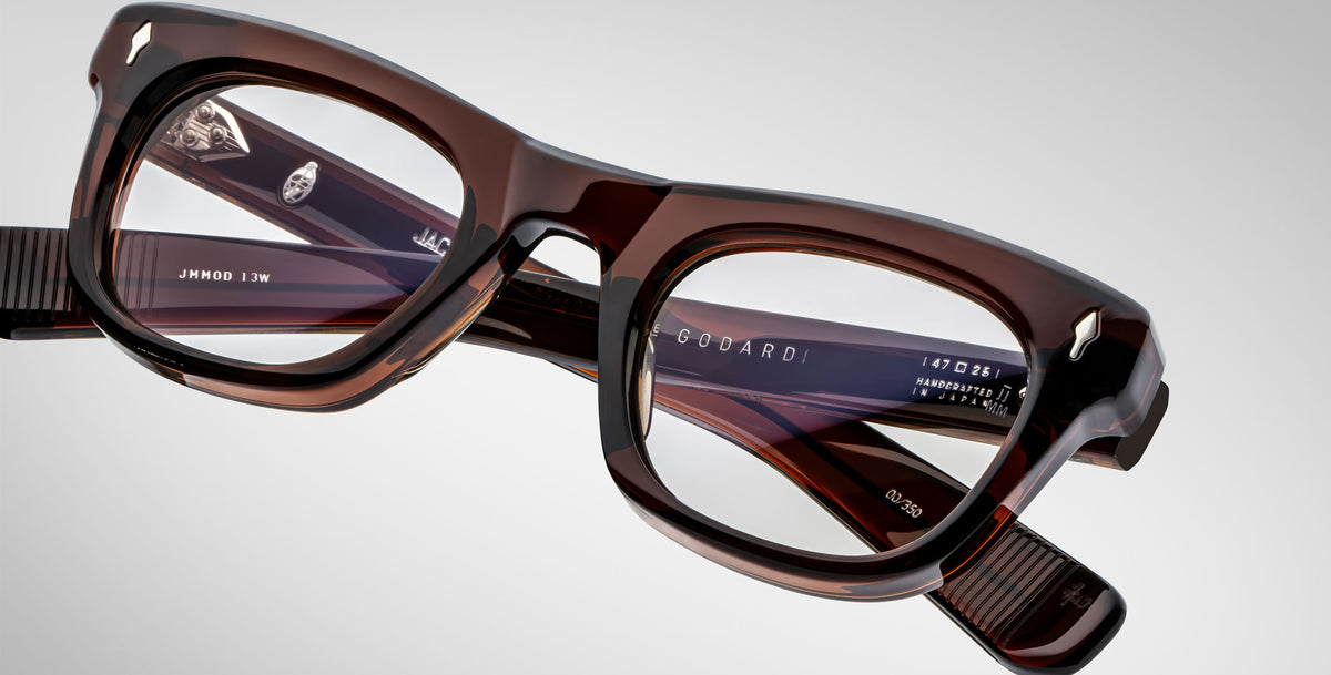 A close-up of rectangular, thick-framed Jacques Marie Mage Godard brown eyeglasses with translucent lenses on a plain gray background. The brand name and model details appear on the inside of the frame arms.