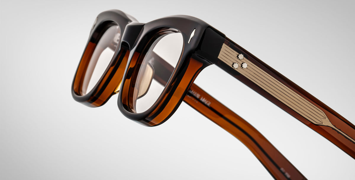 Close-up of Jacques Marie Mage Godard eyeglasses featuring brown and black thick frames, transparent lenses, and metal temple accents, displayed against a plain light background.