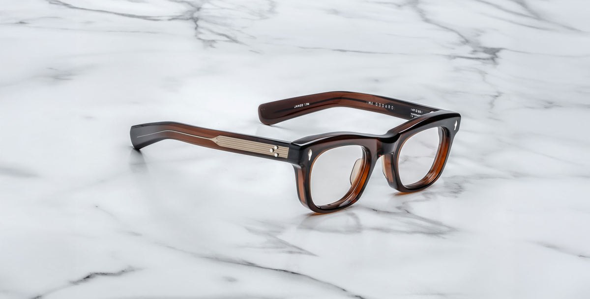 The Jacques Marie Mage Godard eyeglasses feature thick brown tortoiseshell rectangular frames with gold detailing on the temples, displayed on a white marble surface.