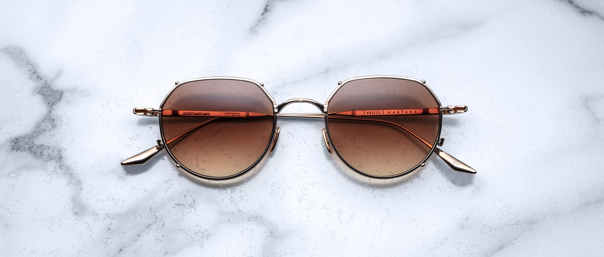 The Jacques Marie Mage Hartana features round, gold frames and brown gradient lenses, shown resting on a white marble surface with gray veining.