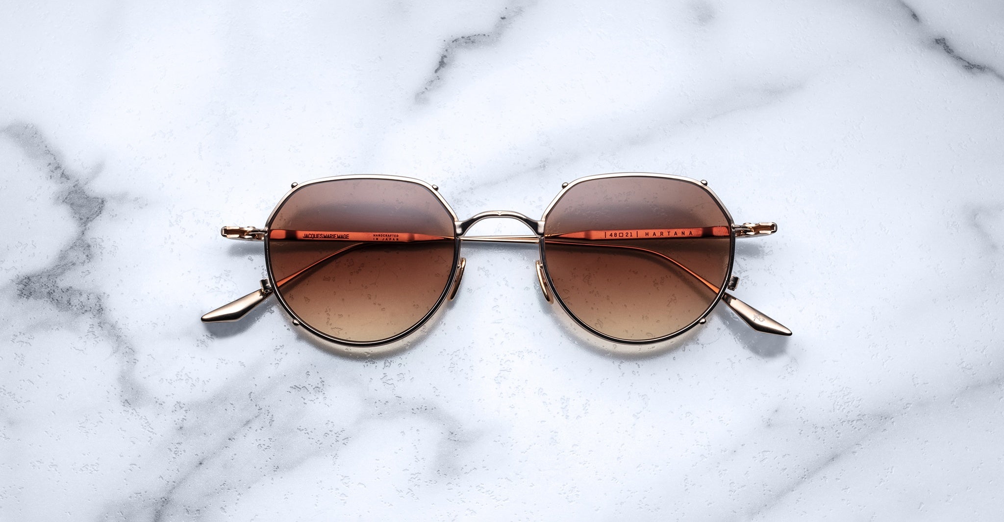 The Jacques Marie Mage Hartana features round, gold frames and brown gradient lenses, shown resting on a white marble surface with gray veining.