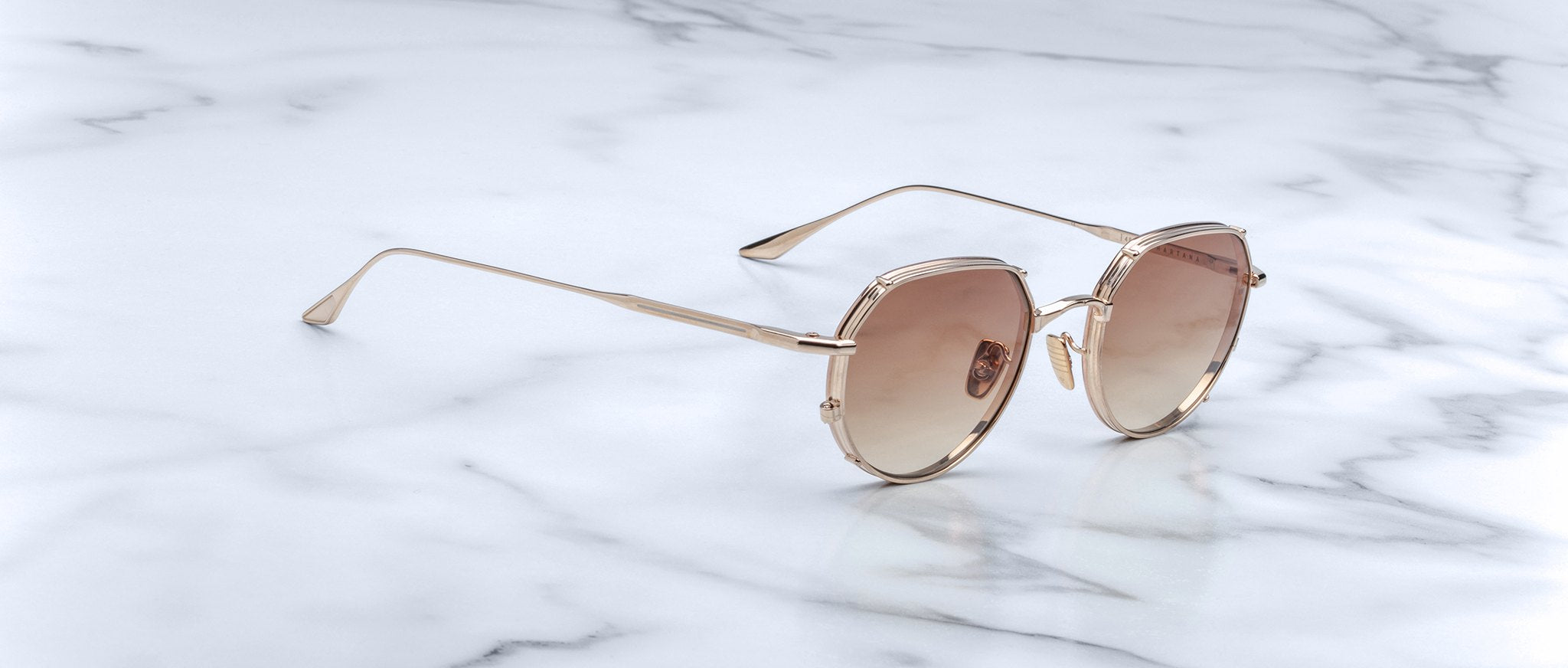 The Jacques Marie Mage Hartana sunglasses feature round, gold frames with light brown tinted lenses and slim metallic temples, displayed on a white marble surface with grey veining.