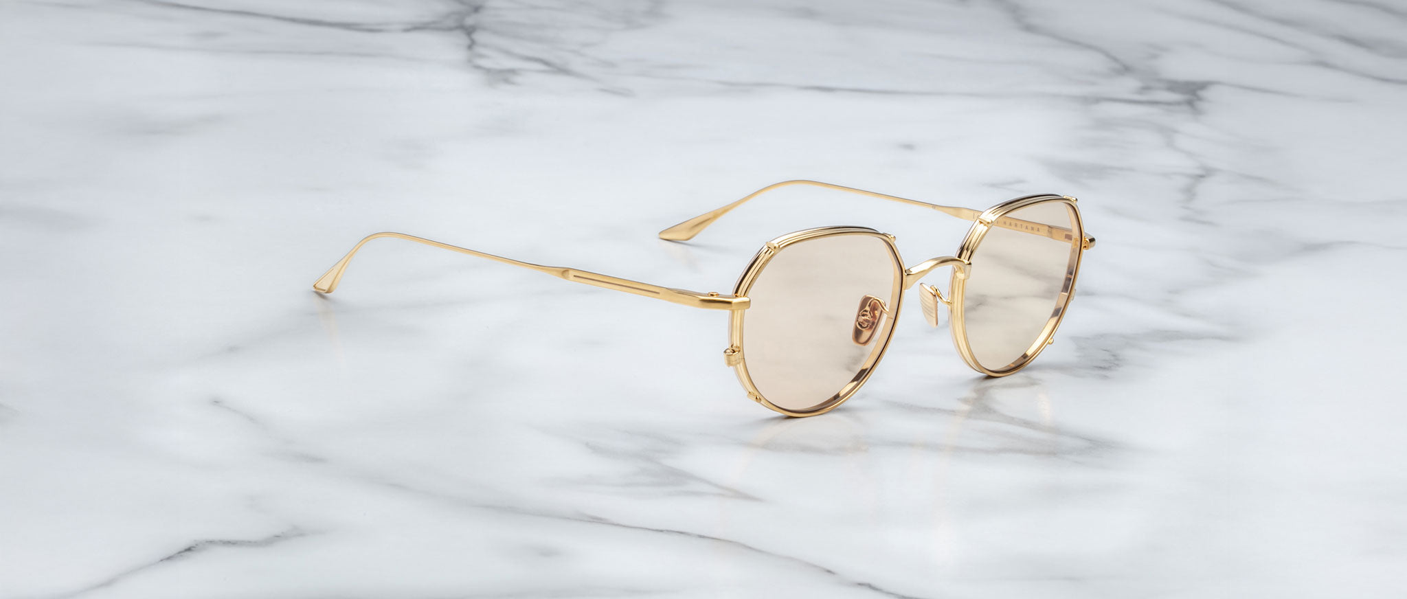 Jacques Marie Mage Hartana gold-framed round sunglasses with lightly tinted lenses rest on a smooth white marble surface with gray veining.