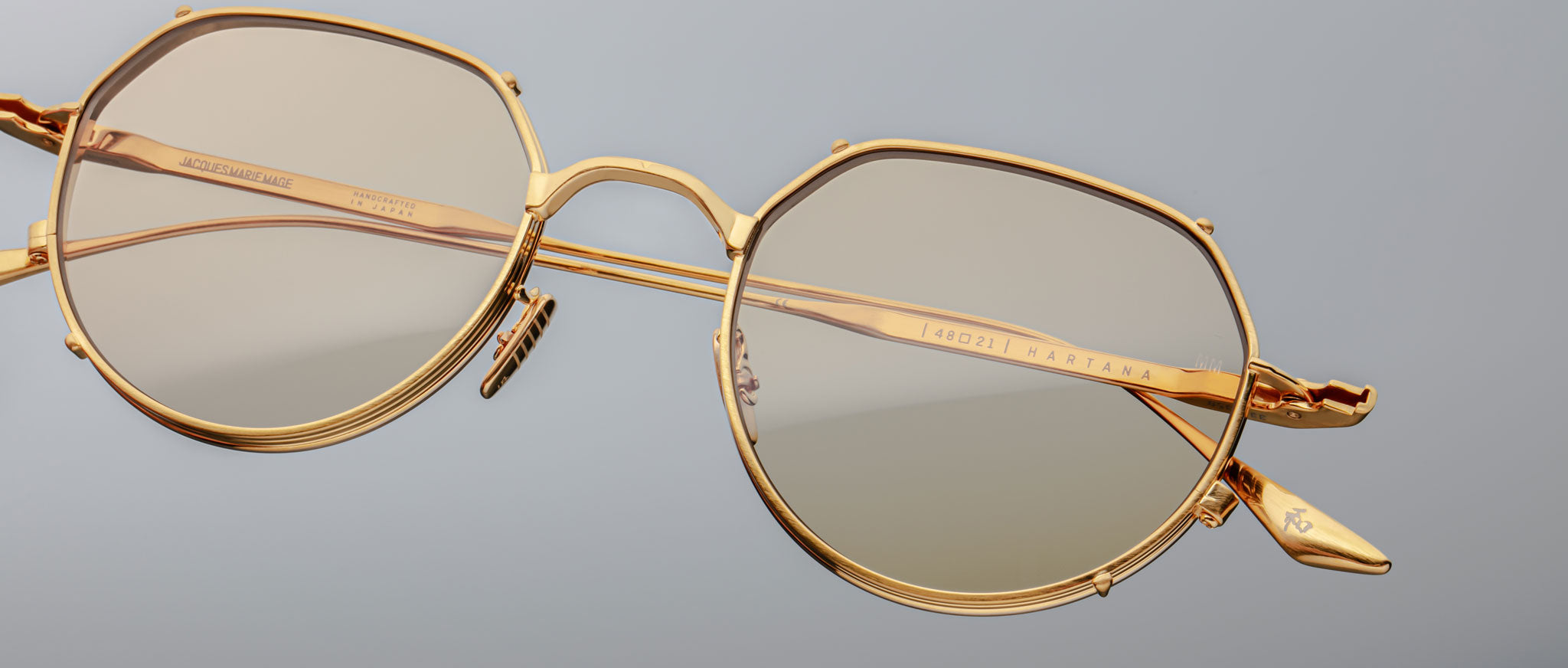 Close-up of Jacques Marie Mage Hartana gold-framed, round eyeglasses with light tinted lenses on a plain light gray background, showcasing fine detailing and visible inscriptions on the arms.
