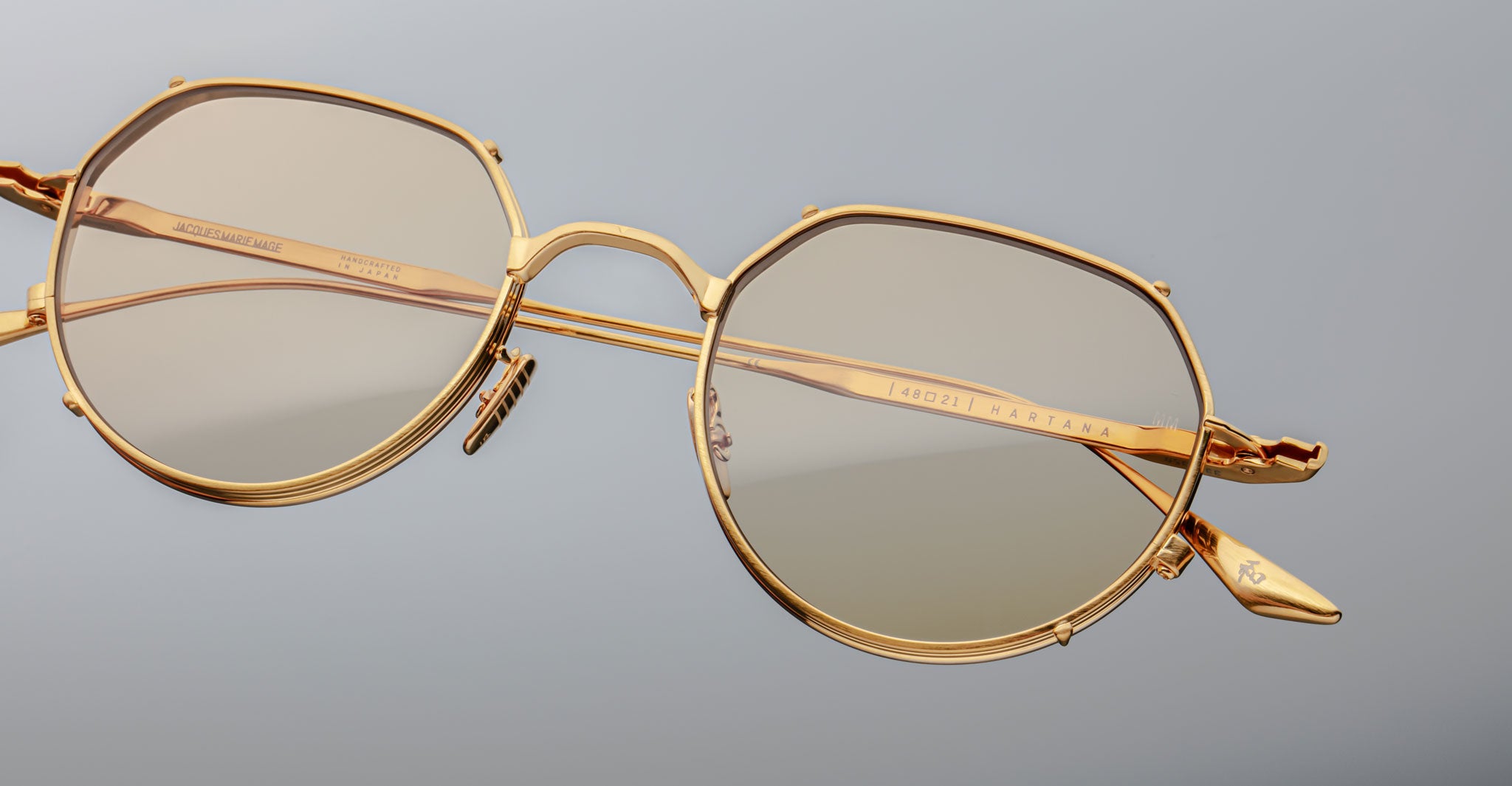 Close-up of Jacques Marie Mage Hartana gold-framed, round eyeglasses with light tinted lenses on a plain light gray background, showcasing fine detailing and visible inscriptions on the arms.