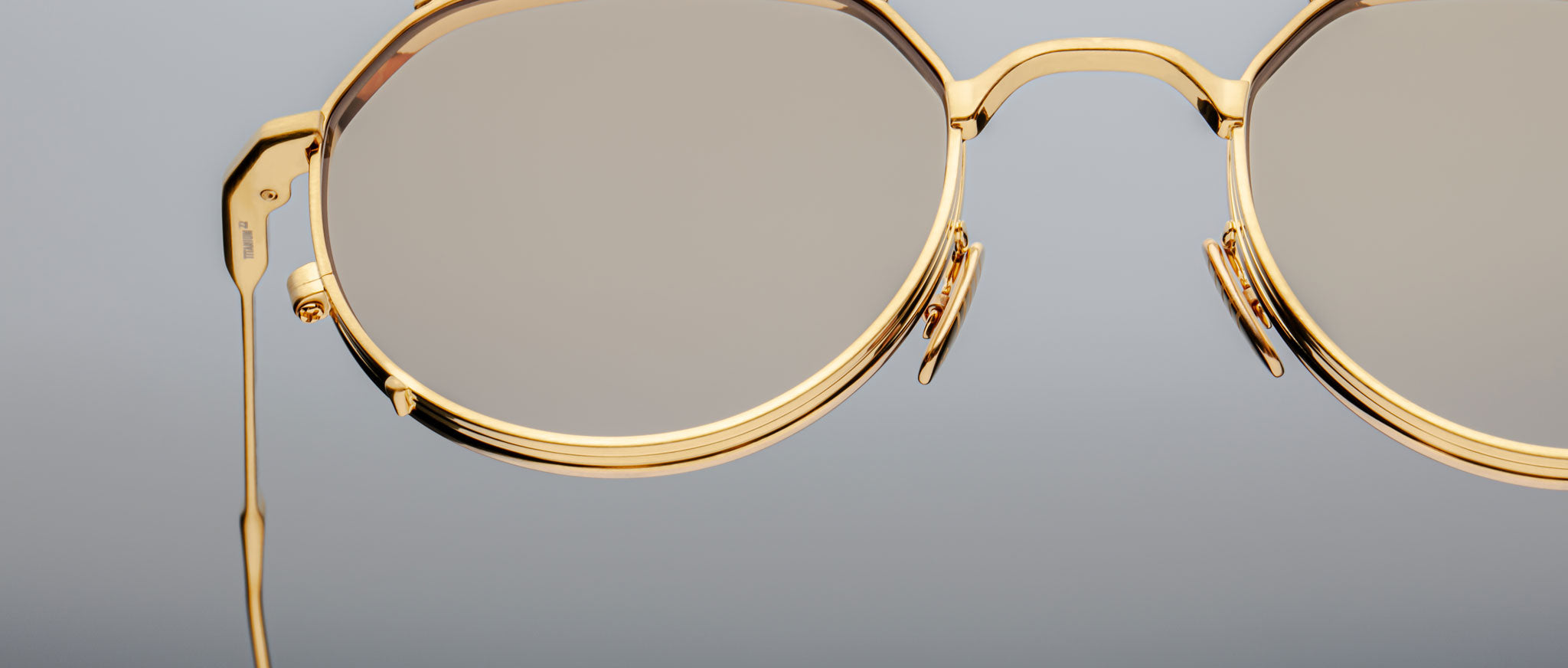Close-up of Jacques Marie Mage Hartana eyeglasses with stylish gold frames and round lenses, set against a plain gray background. Only the right half of the glasses is visible in the image.
