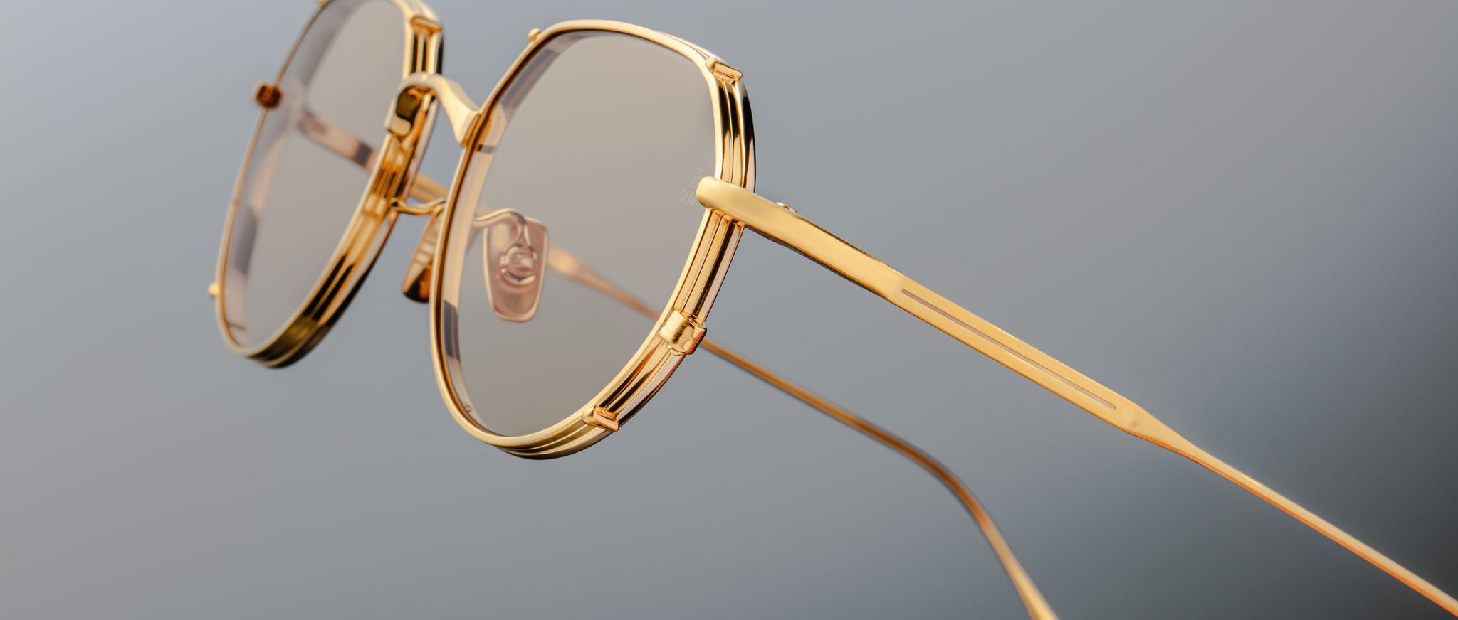 Close-up of Jacques Marie Mage Hartana eyeglasses featuring stylish gold frames, round lenses with a light brown tint, and thin temples, set against a plain grey background.