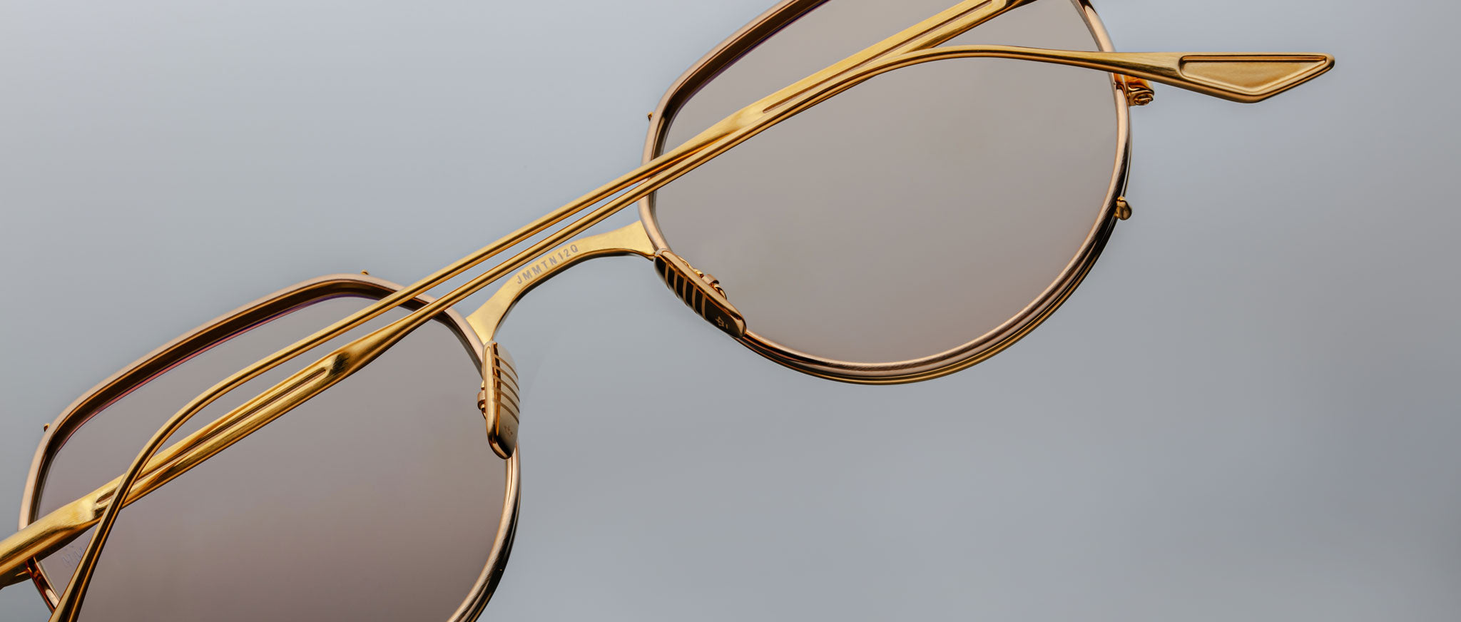 Close-up of Jacques Marie Mage Hartana sunglasses with stylish gold frames and brown tinted lenses, displayed on a plain gray background. The image showcases the sleek metal frame and modern design details.