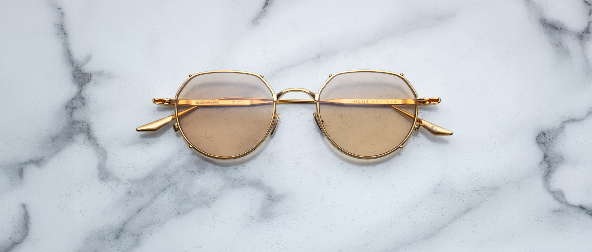 The Jacques Marie Mage Hartana features gold frames and round brown-tinted lenses, displayed on a white marble surface with grey veining.