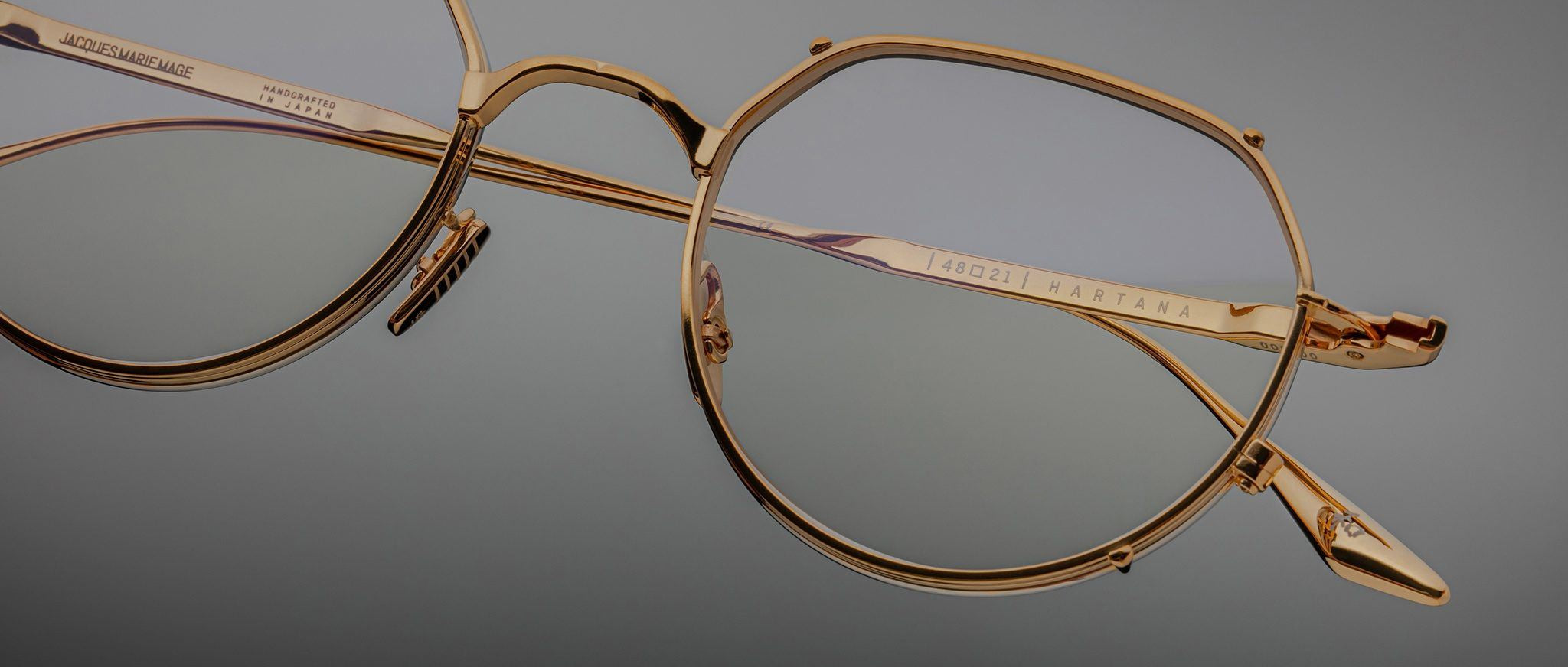 Close-up of Jacques Marie Mage Hartana gold metal eyeglass frames with clear lenses, detailed hinges, and engraved text on the arms, set against a neutral gray background.