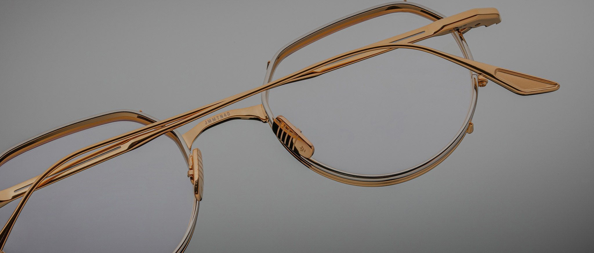 Close-up of Jacques Marie Mage Hartana gold-framed eyeglasses with clear lenses on a neutral gray background, highlighting the frame’s fine details and craftsmanship.