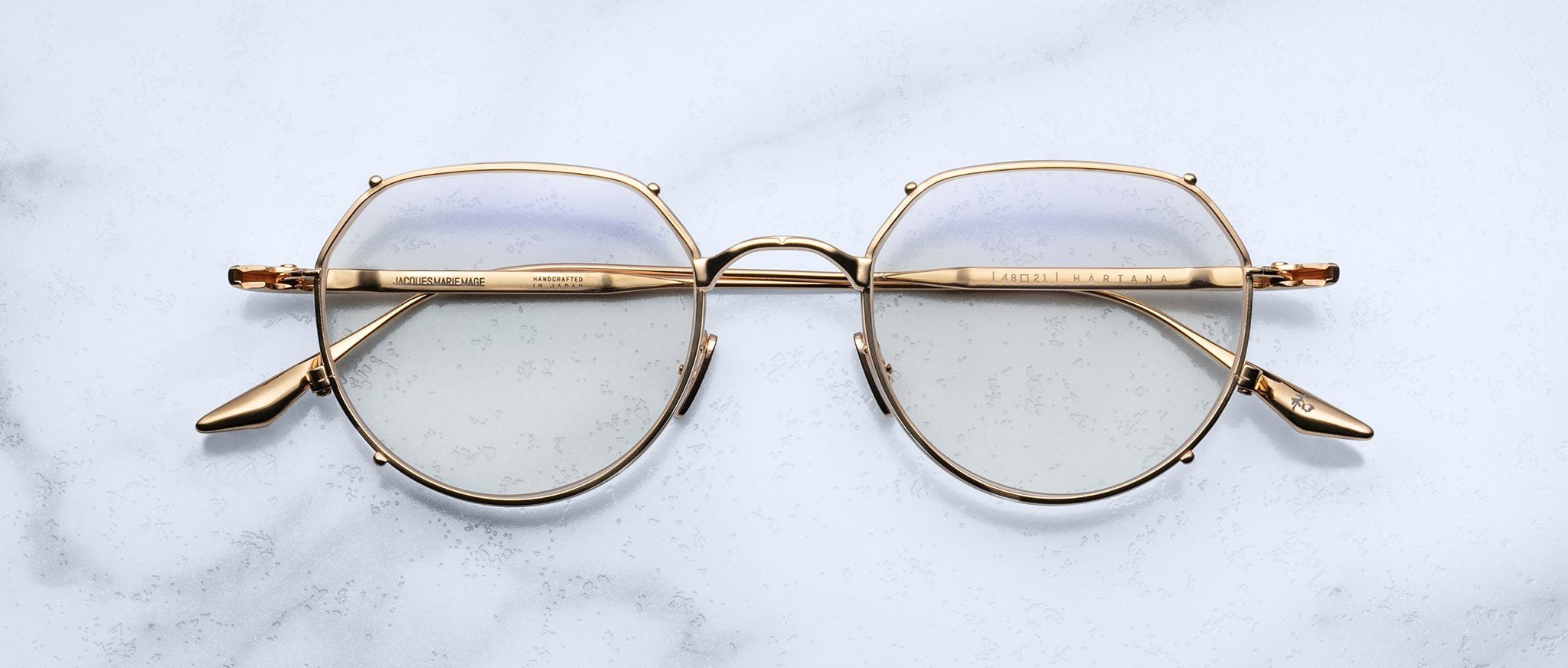 The Jacques Marie Mage Hartana gold metal round eyeglasses with clear lenses rest on a white marble surface, featuring subtly engraved arms and visible nose pads.