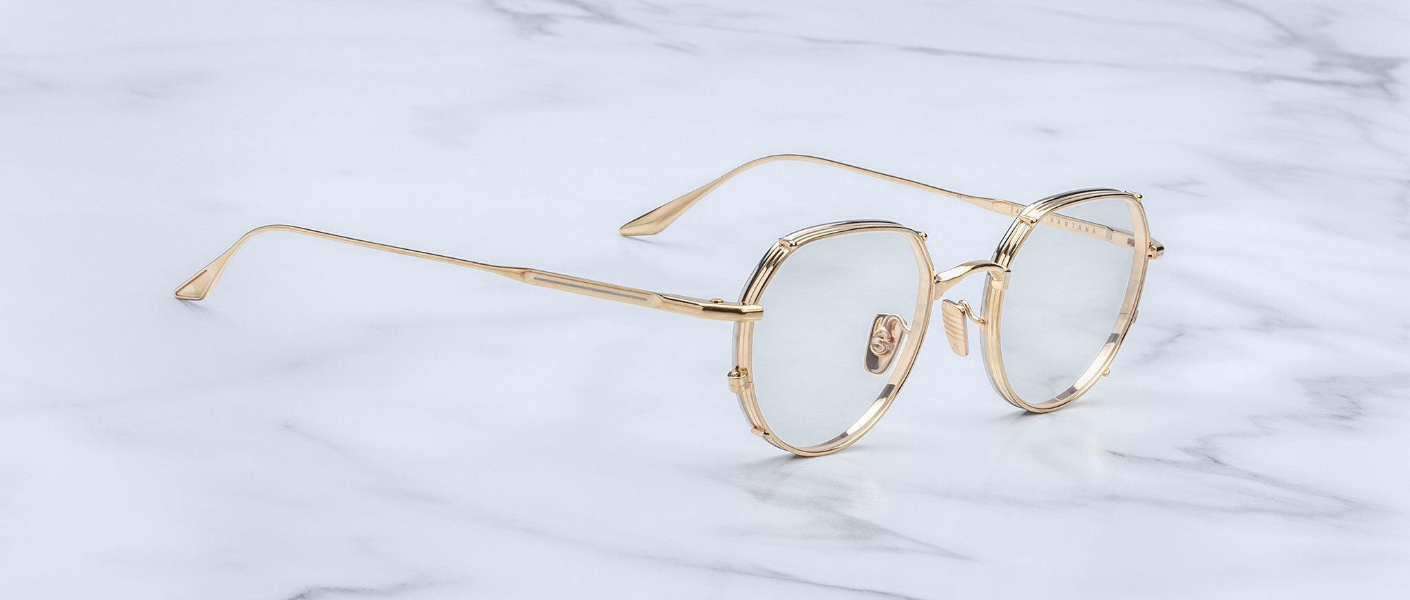 Jacques Marie Mage Hartana eyeglasses with a gold metal frame and clear lenses are displayed on a white marble surface.