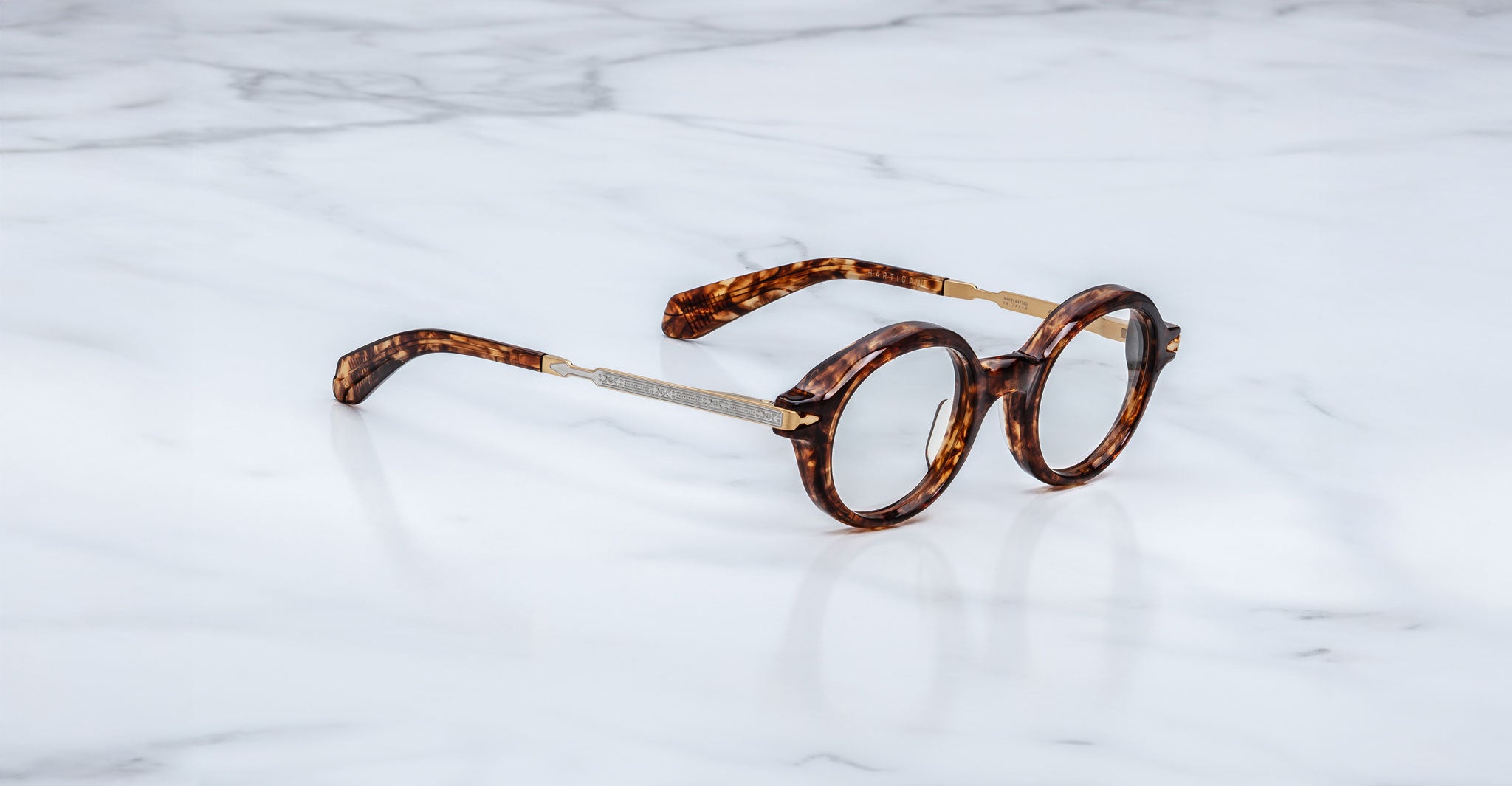 Jacques Marie Mage Hartigan in Argyle, round tortoiseshell acetate eyeglasses shown from a side angle, highlighting engraved metal core temples, keyhole bridge, and sculpted temple tips on a light marble surface.v