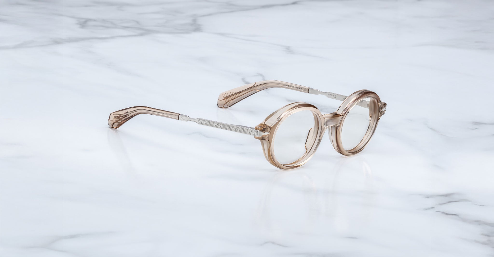 Jacques Marie Mage Hartigan in Sand, round translucent beige acetate eyeglasses shown from a side angle, highlighting engraved metal core temples, keyhole bridge, and sculpted temple tips on a light marble surface.