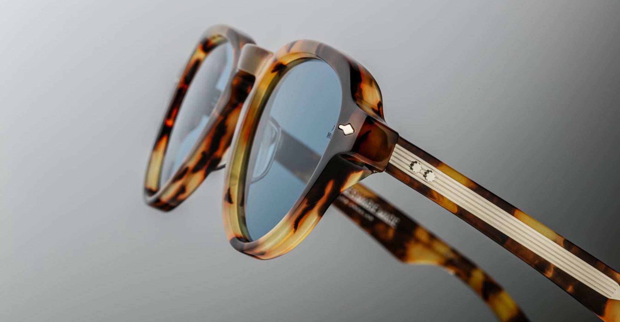 Close-up of Jacques Marie Mage Hatfield tortoise shell sunglasses featuring blue-tinted lenses, silver hinge details, and textured metal-accented temples set against a neutral gradient background.