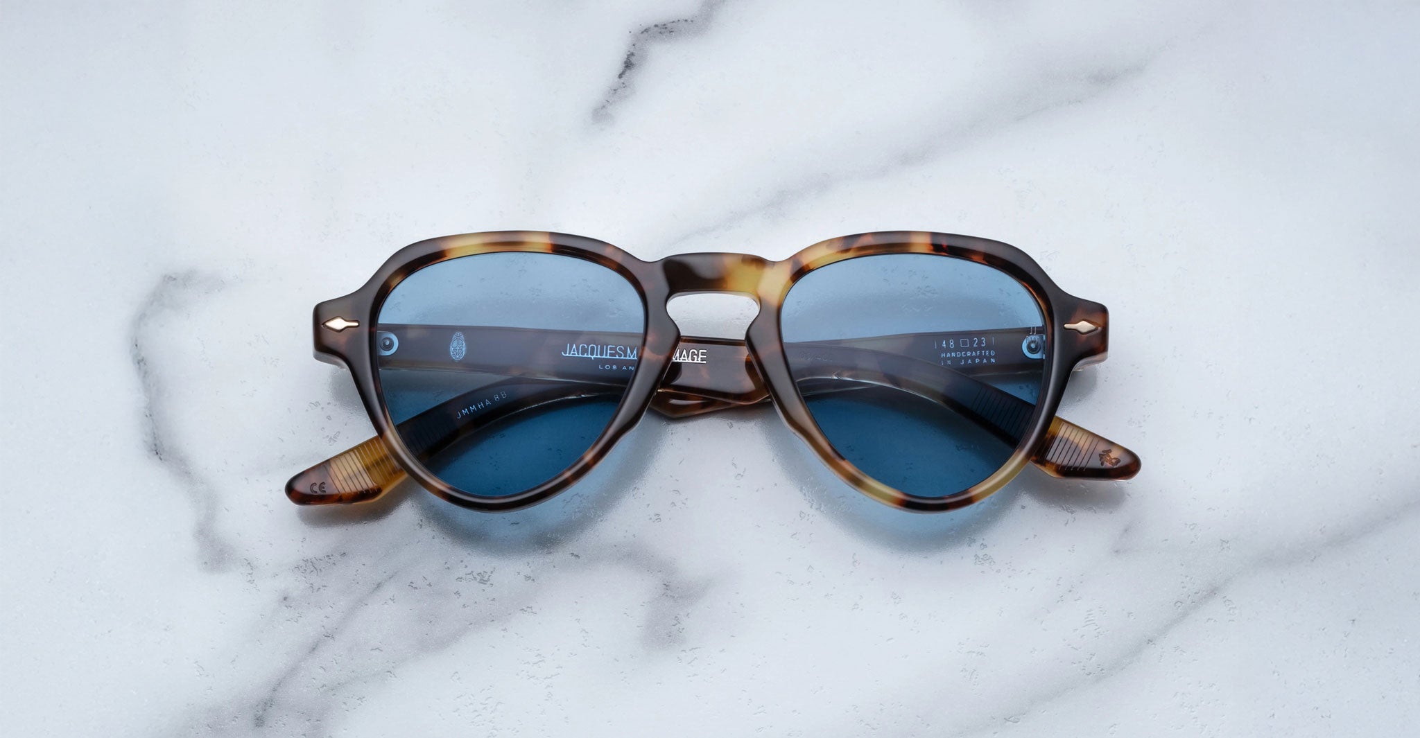 The Jacques Marie Mage Hatfield sunglasses, featuring brown tortoiseshell frames and blue-tinted lenses, are displayed on a white marble surface.
