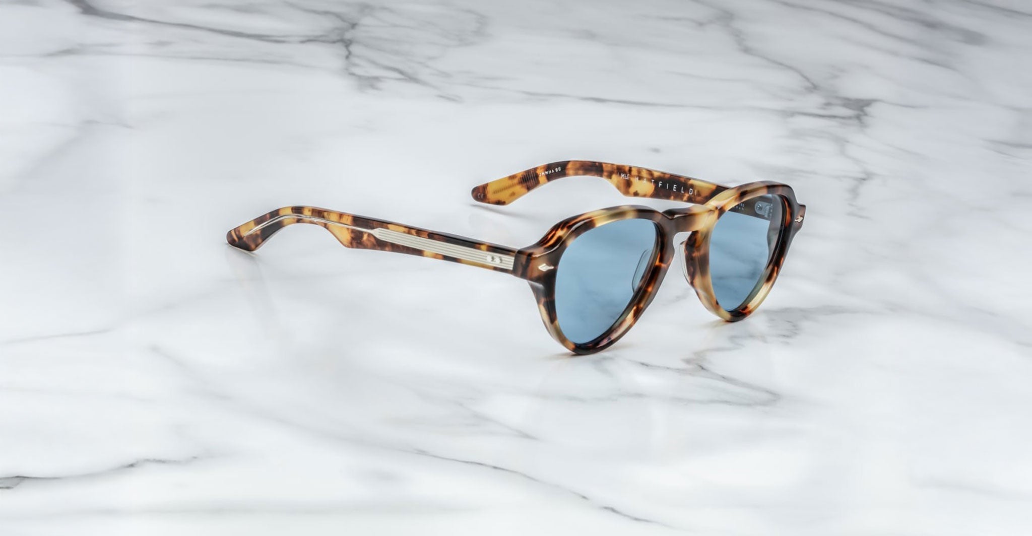 The Jacques Marie Mage Hatfield sunglasses, featuring a tortoiseshell pattern and blue tinted lenses, rest on a white marble surface.