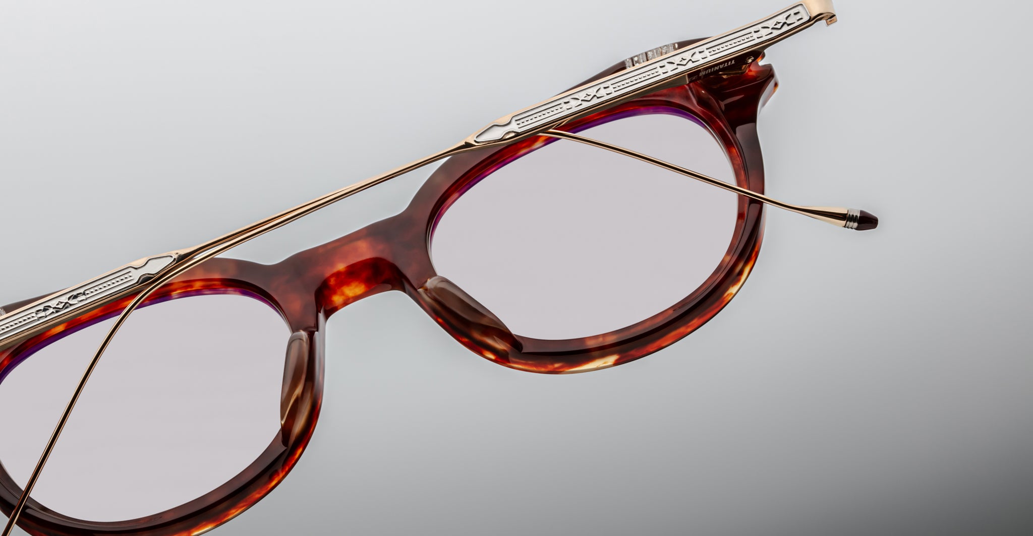 Close-up of Jacques Marie Mage Hisao tortoiseshell eyeglasses with decorative gold-colored metal arms resting diagonally over the lenses, set against a light grey background.
