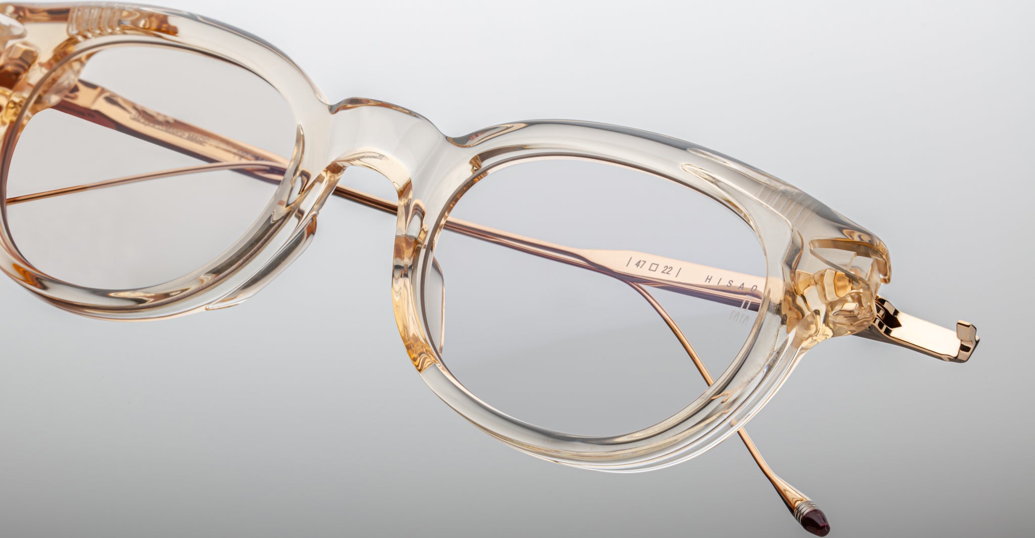 The Jacques Marie Mage Hisao features clear, round eyeglasses with transparent beige frames and thin gold temples, shown against a light gray background.