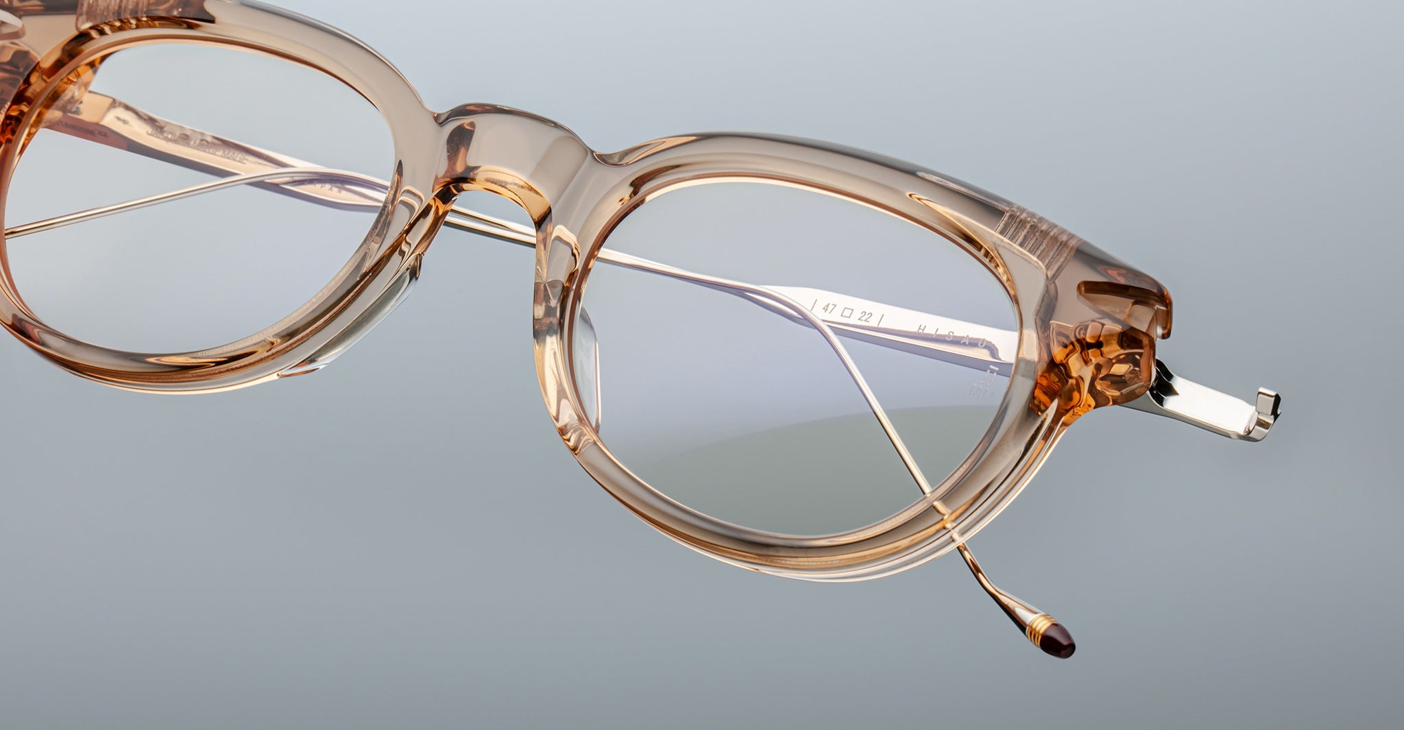 A close-up of Jacques Marie Mage Hisao eyeglasses featuring clear light brown round frames, displayed at an angle against a plain, neutral background.